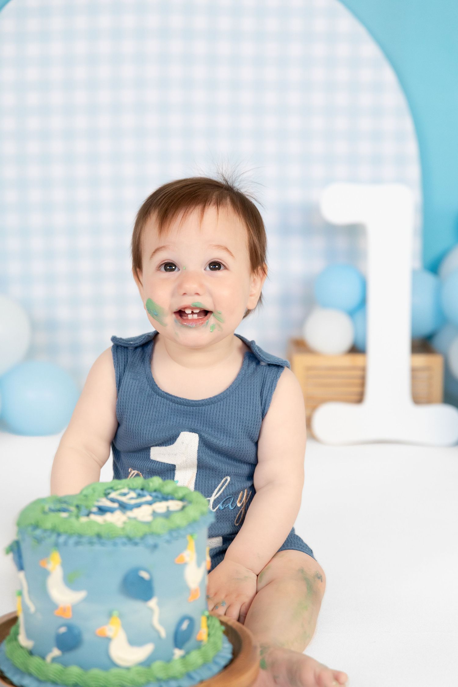 Baby boy celebrates first birthday smashing blue duck-themed cake, face covered in green frosting, wearing number 1 romper.