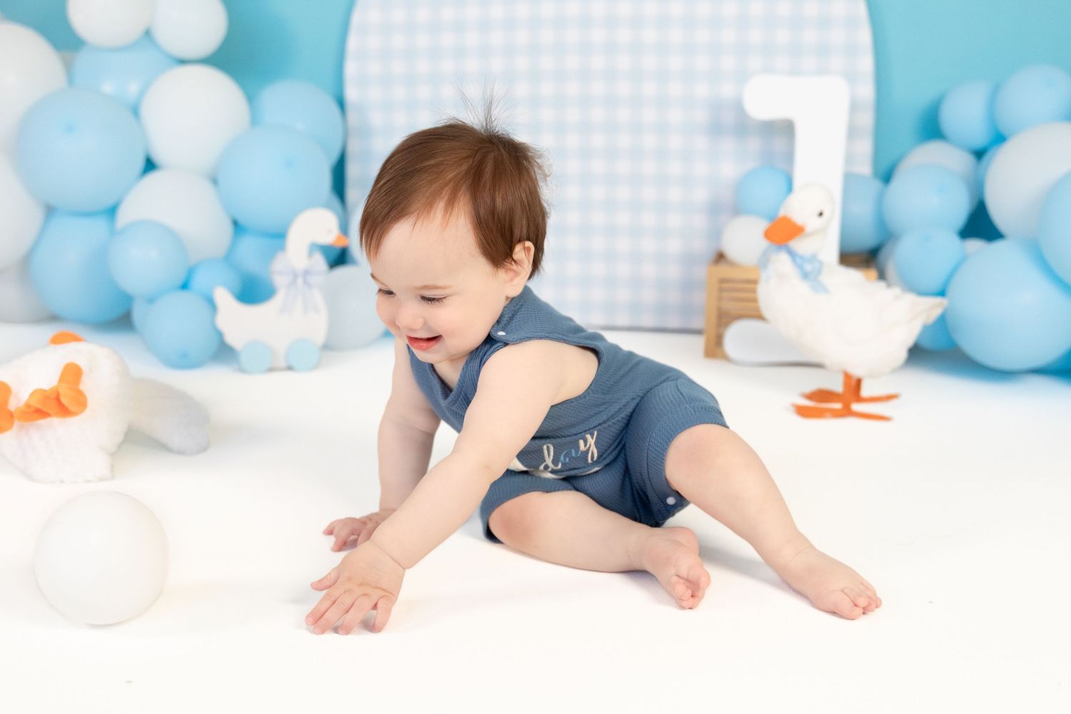 Laughing baby boy in denim romper crawls on white floor surrounded by blue balloons and duck decorations.