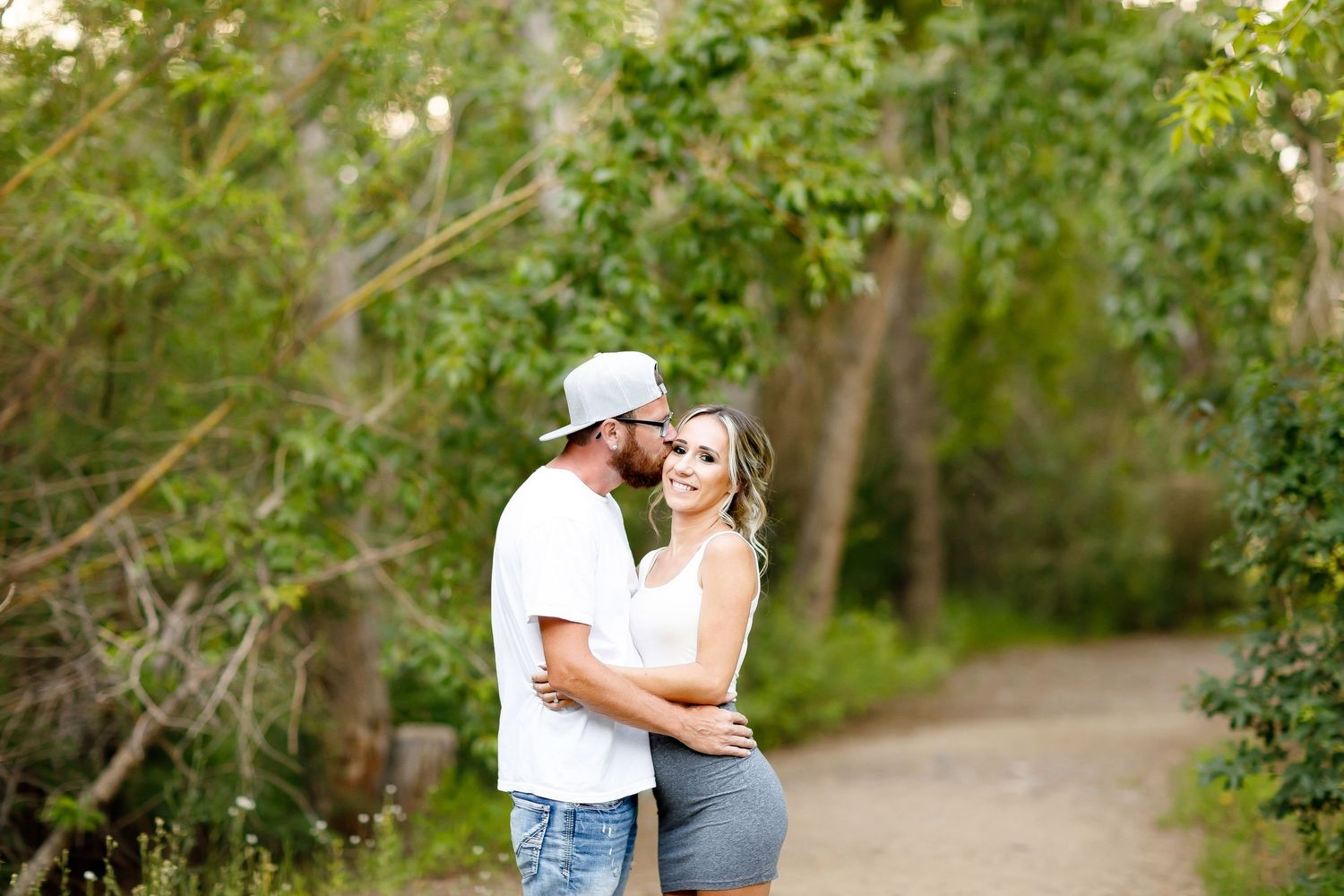 Couple embracing and kissing on a garden path surrounded by lush green trees during an outdoor engagement session.