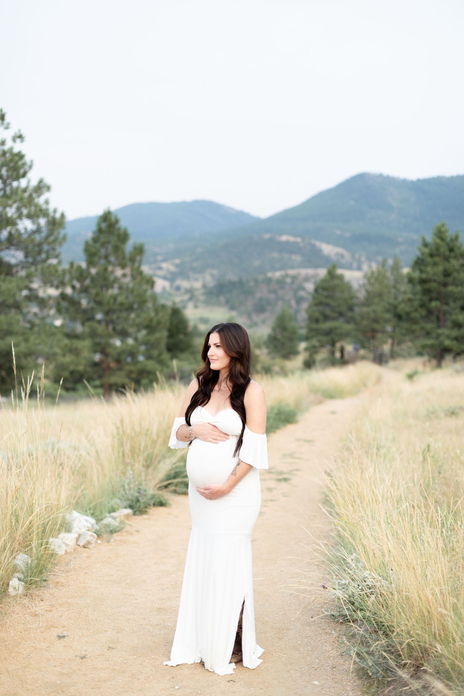 Pregnant woman in elegant white off-shoulder gown stands on a mountain path cradling her belly.