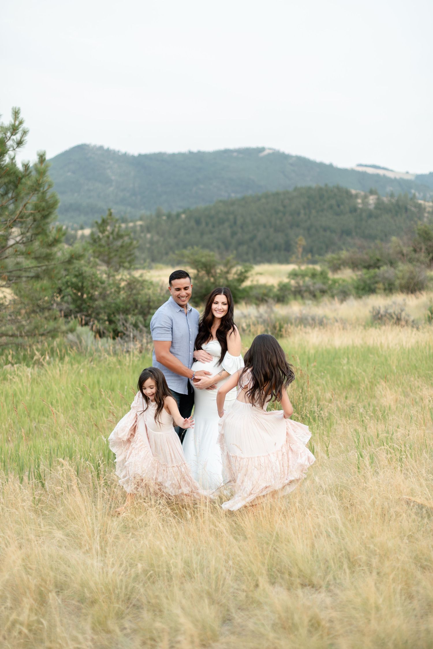 Family of four in mountain meadow with girls twirling in pink dresses.