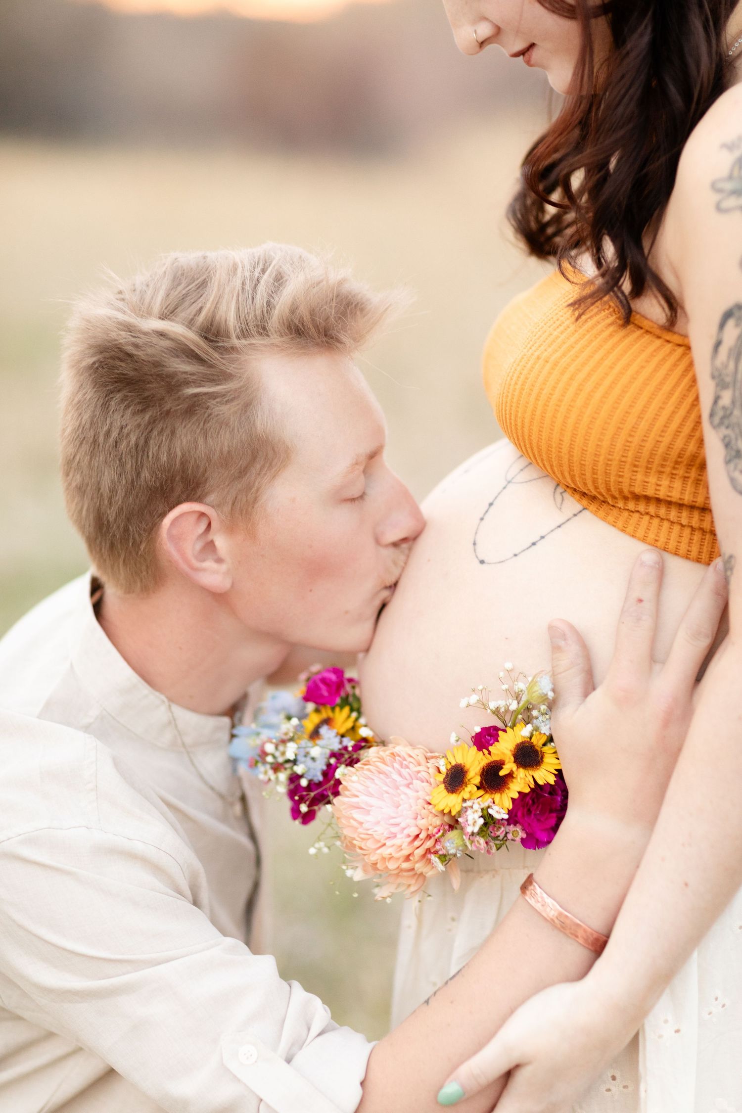 Close-up of man kissing pregnant woman's bare belly while holding colorful sunflower bouquet outdoors.