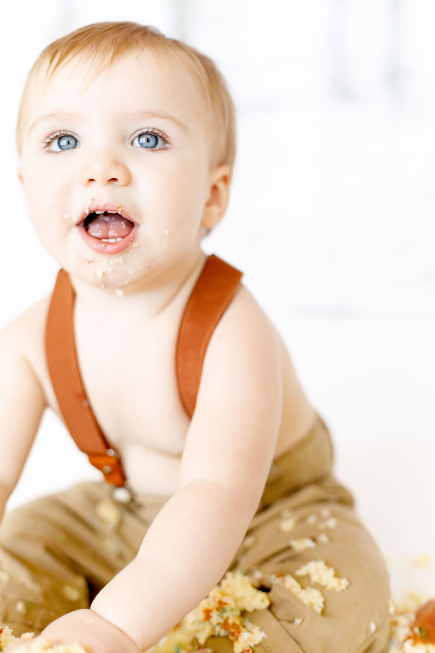Happy blue-eyed baby boy with cake on face wearing suspenders during first birthday smash.