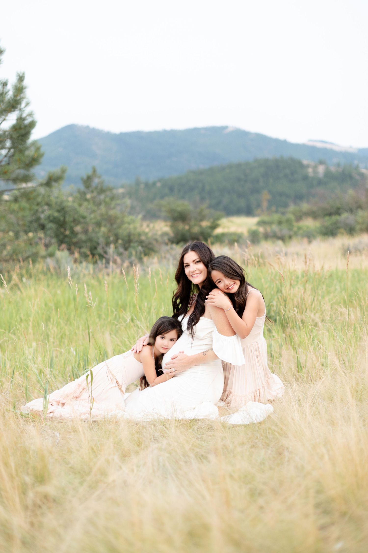 Pregnant mother sitting in tall grass with two daughters embracing her.