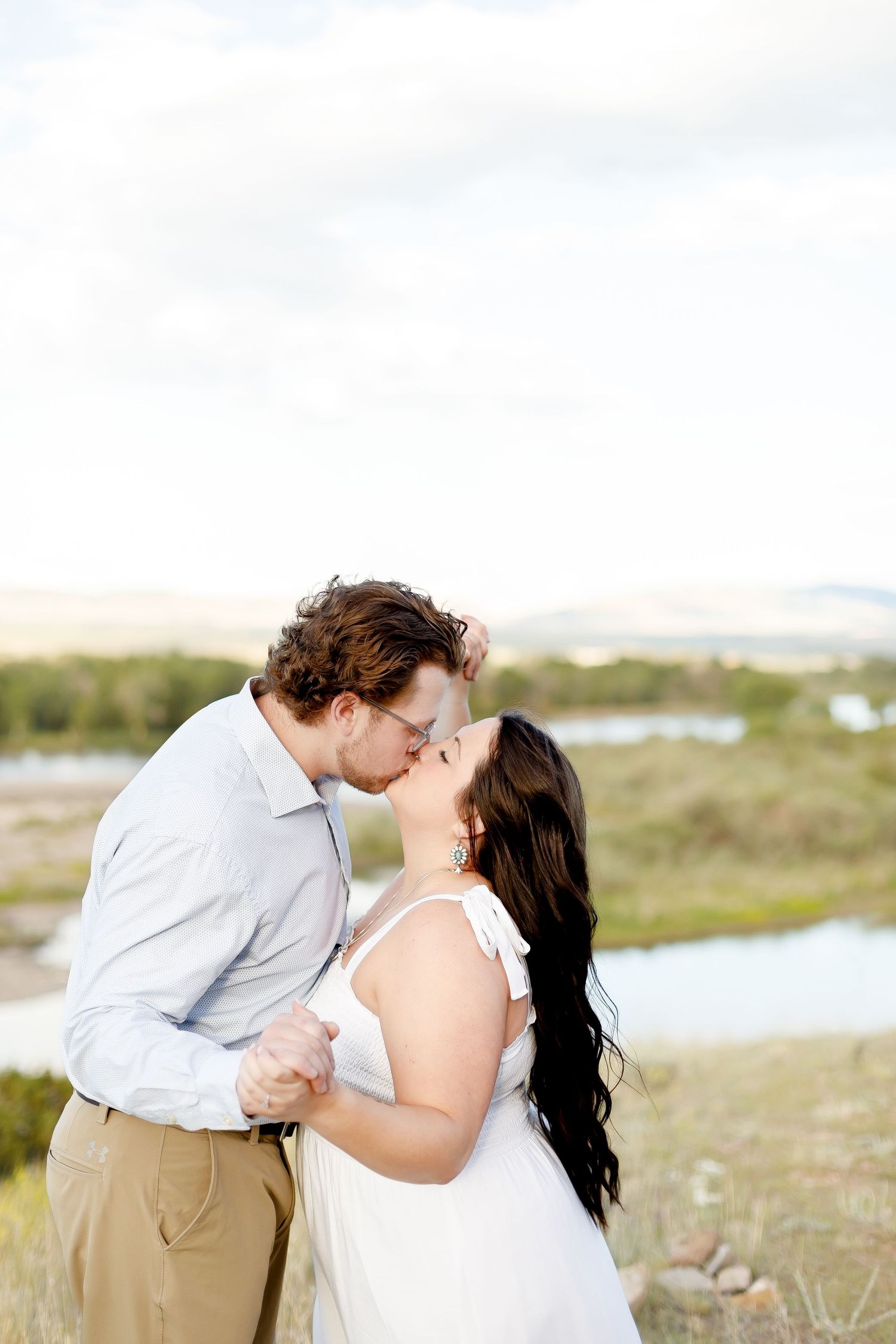 Couple sharing a kiss near a scenic lake with mountains, woman in white dress with long dark hair.