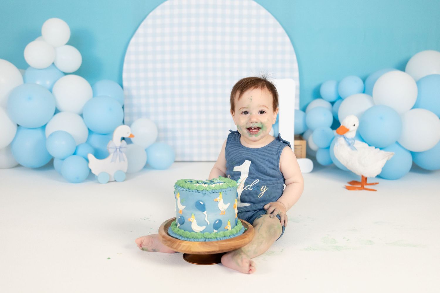 Happy baby boy sits with blue duck-decorated first birthday cake surrounded by blue balloons and duck props.