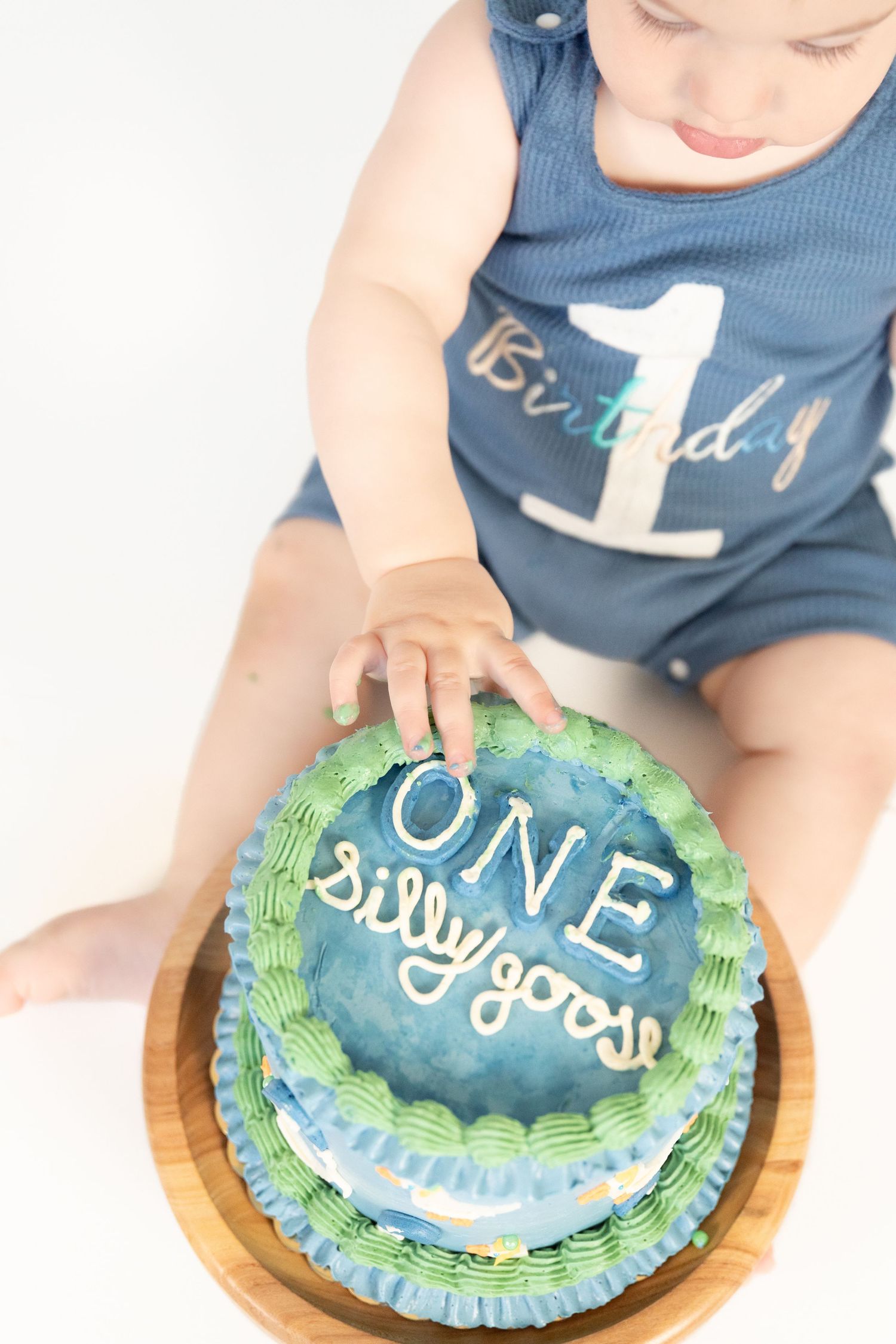 Overhead view of baby boy in birthday romper reaching into blue and green ONE silly goose smash cake.
