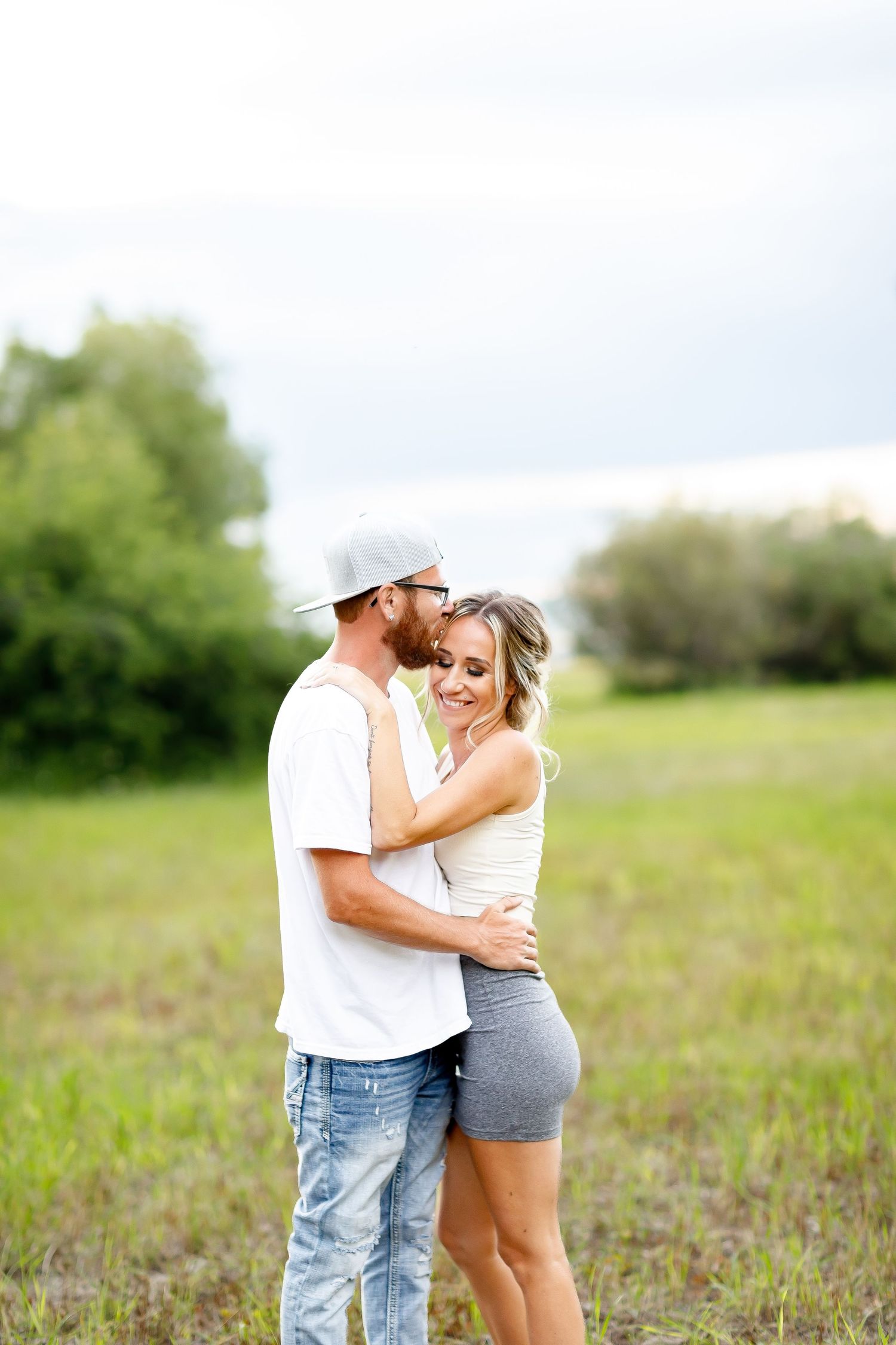 Man in white tee and cap kissing woman in white top and gray shorts while embracing in a green field.
