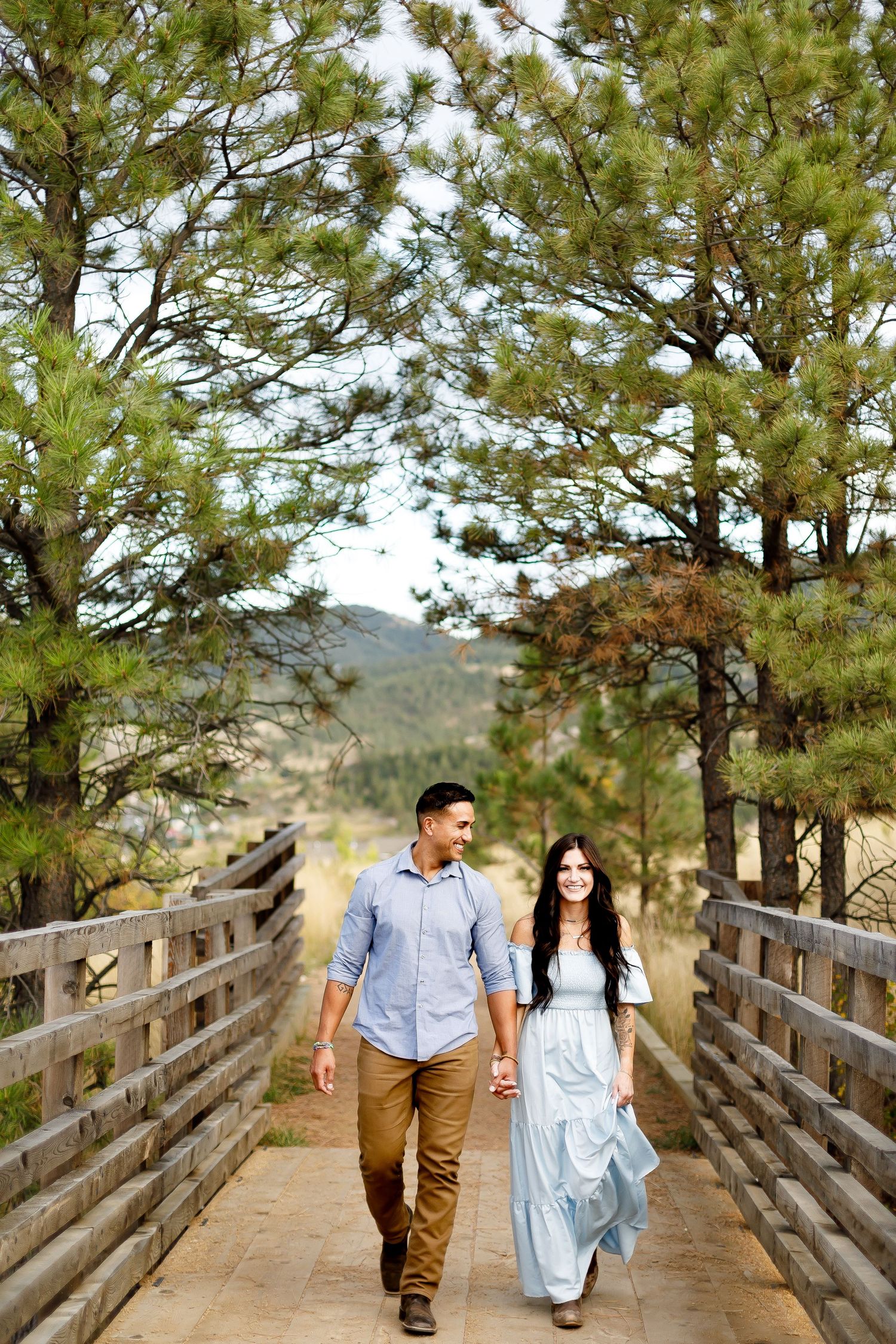 Couple holding hands and walking across a wooden bridge surrounded by pine trees during a mountain engagement session.