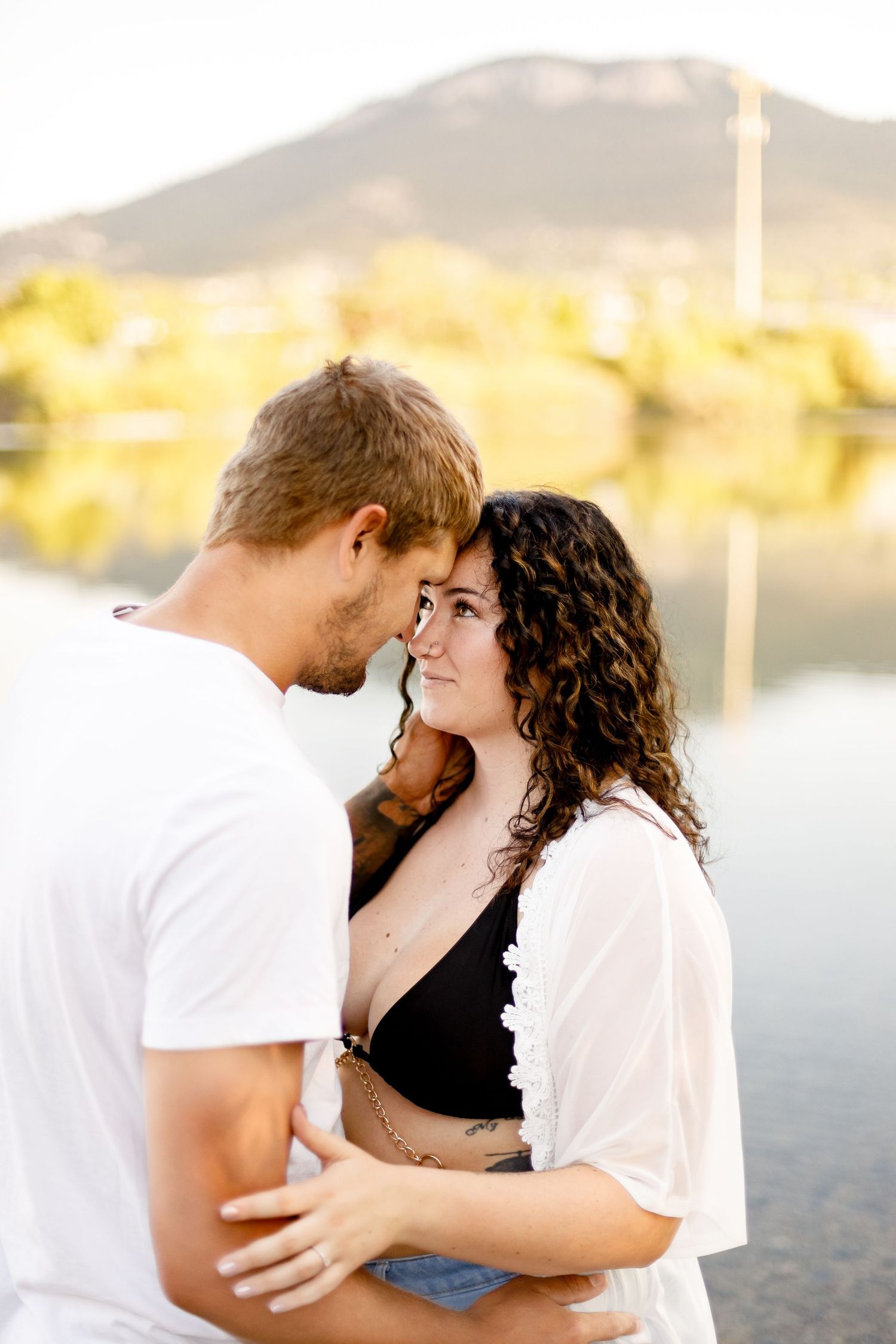 Romantic couple touching foreheads while embracing near a lake with mountain backdrop at sunset.