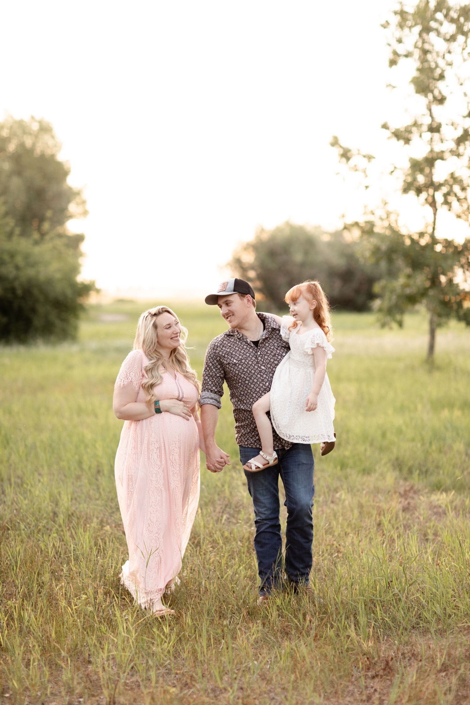 Family maternity photo with pregnant woman in pink dress, man in cowboy hat holding young redhead girl.