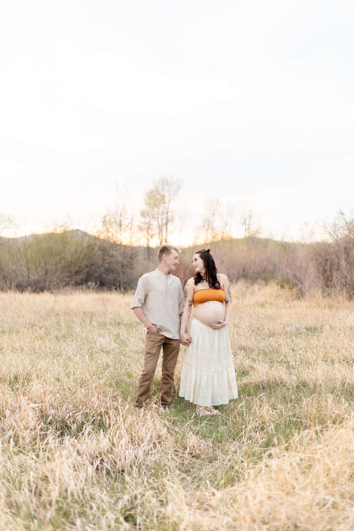 Pregnant couple holds hands smiling at each other in golden field at sunset, woman in orange crop top.