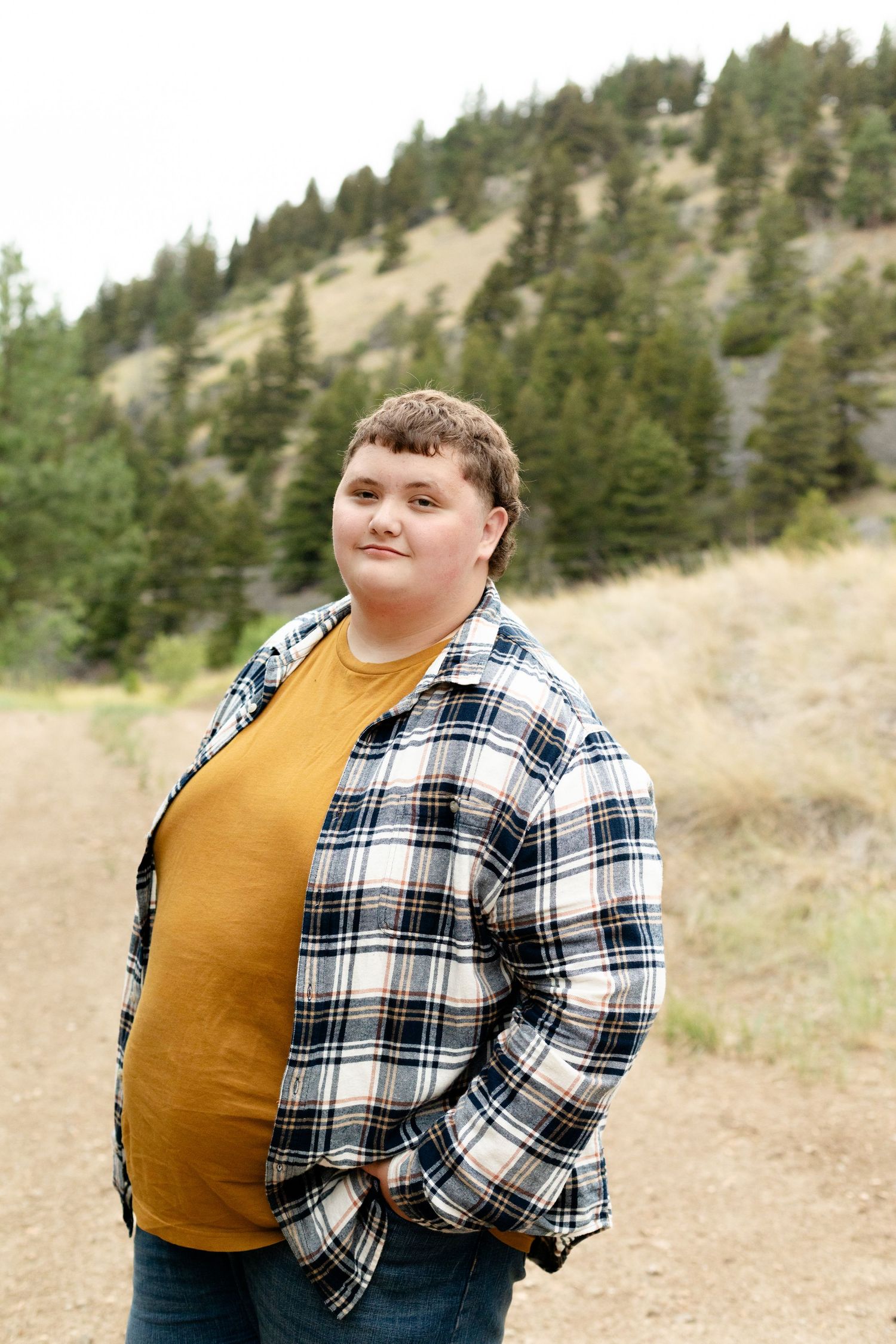 Young person in plaid shirt and mustard tee standing on a dirt trail with pine-covered hills behind them.