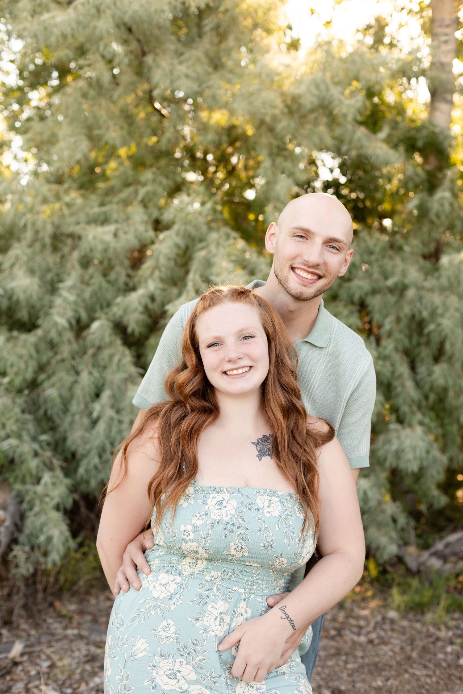 Smiling couple posing together in front of green trees, woman in floral dress outdoors.
