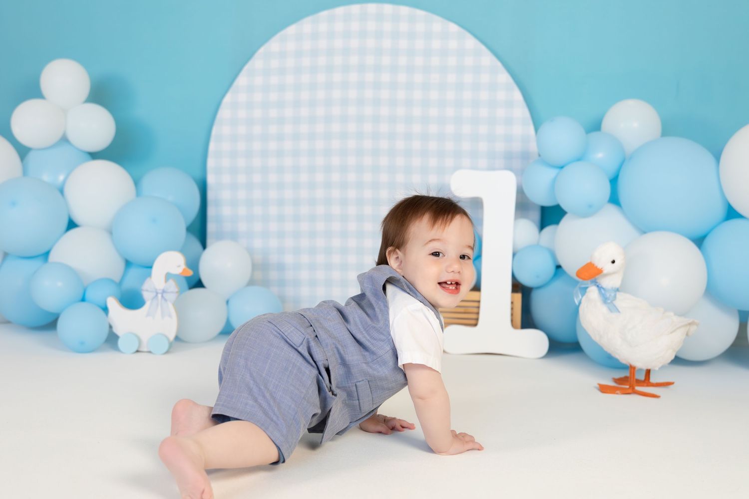 Smiling baby boy crawls toward camera during first birthday photo session with blue balloons and duck decor.