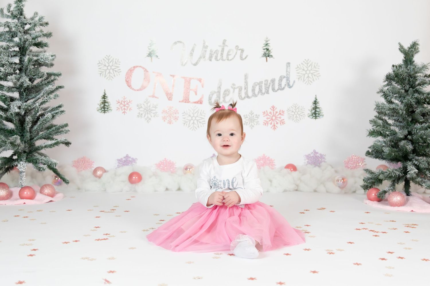 Baby girl in pink tutu sitting before Winter ONEderland first birthday backdrop with Christmas trees.