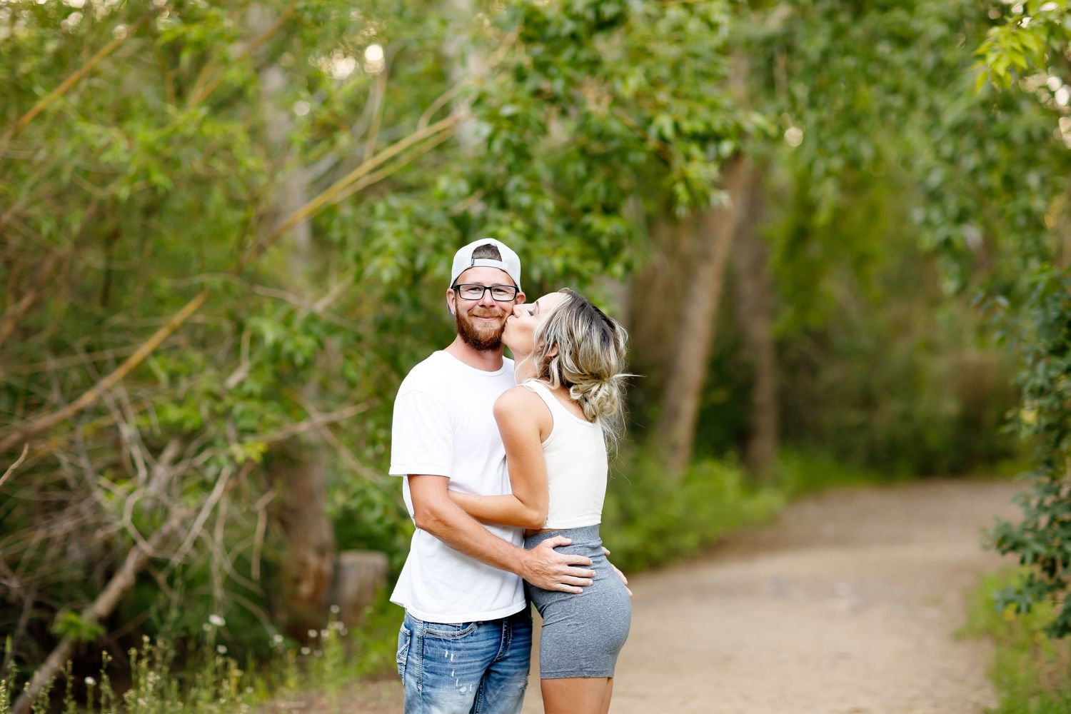 Woman kissing mans cheek as they embrace on a lush green tree-lined garden path.