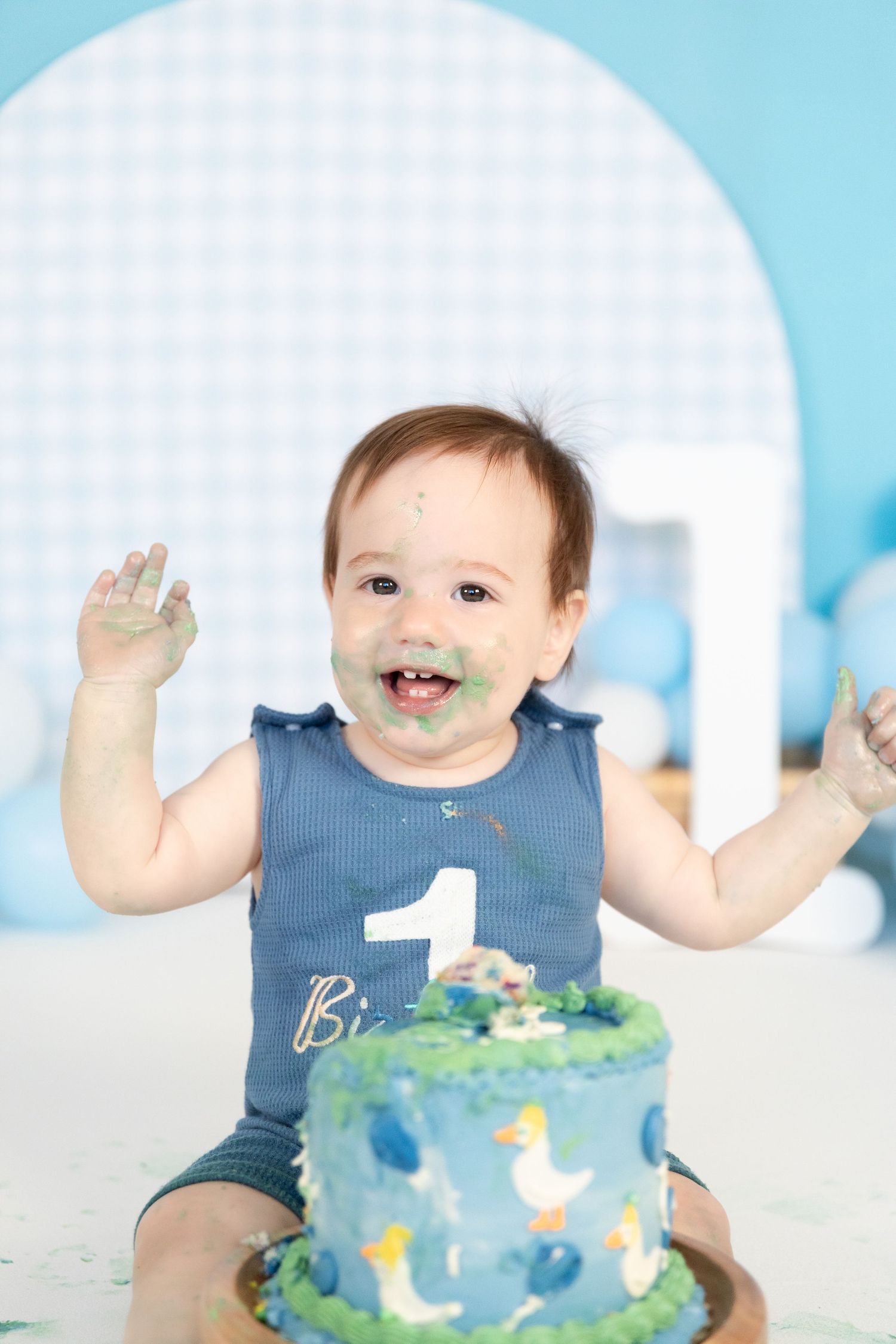Happy baby boy celebrating first birthday with smashed blue farm animal cake, hands raised joyfully.