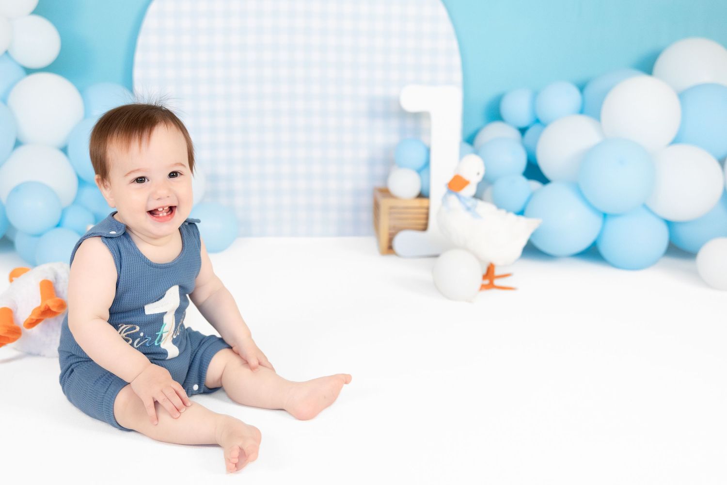 Laughing baby boy in a blue birthday romper sitting on white floor surrounded by blue balloons.