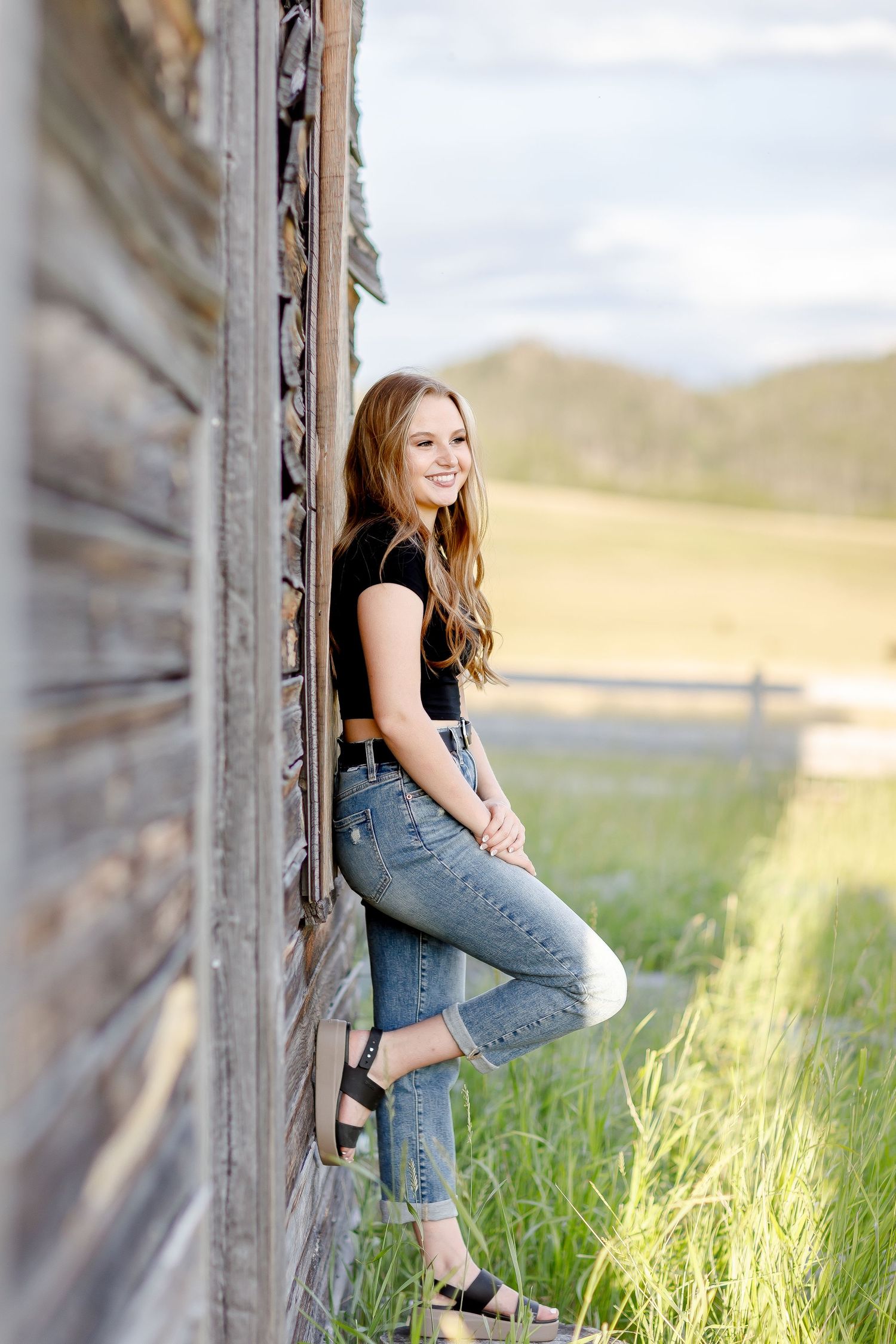 Teen girl in black crop top and jeans leaning against a weathered barn wall in a green field.