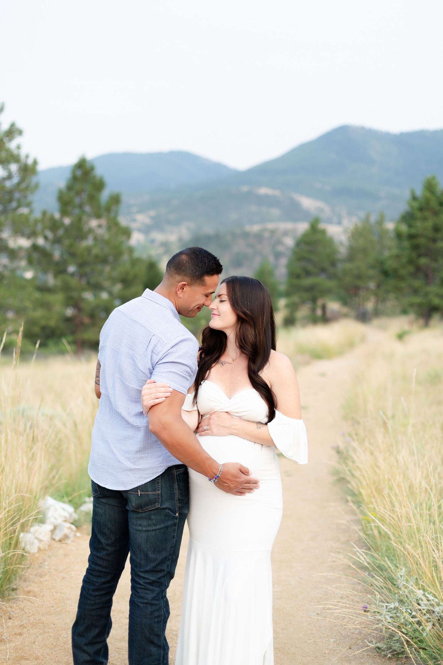 Couple embracing on a scenic mountain trail, man kissing woman's head as she smiles in white gown.
