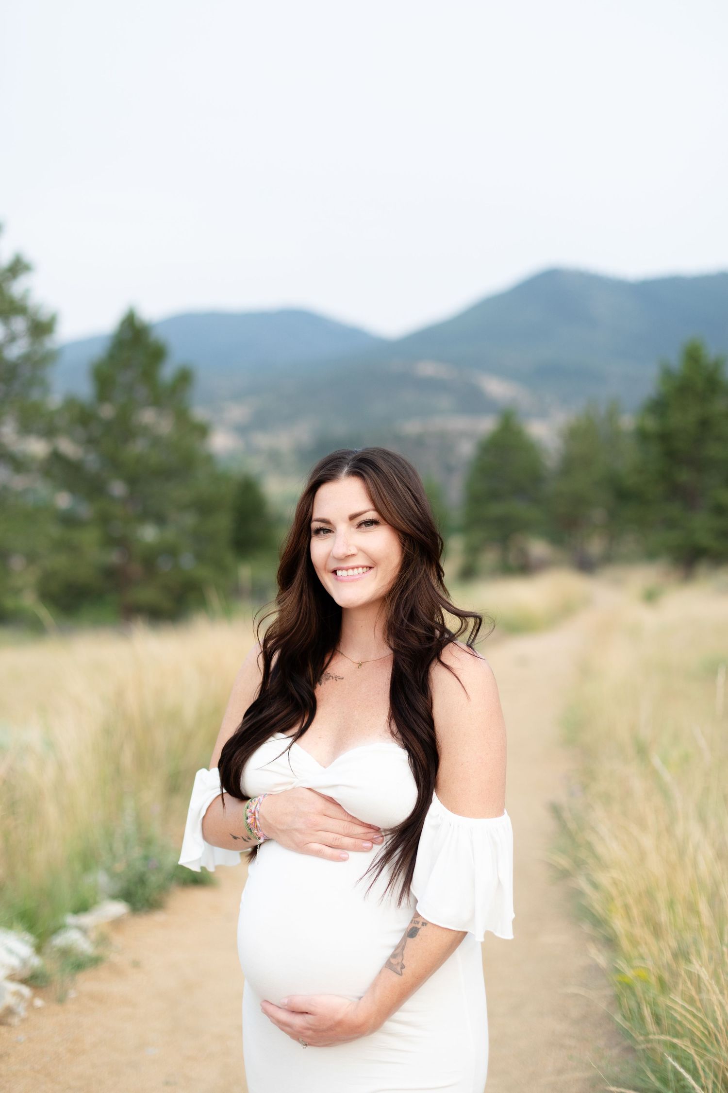 Smiling pregnant woman in white off-shoulder dress holding baby bump on mountain trail.