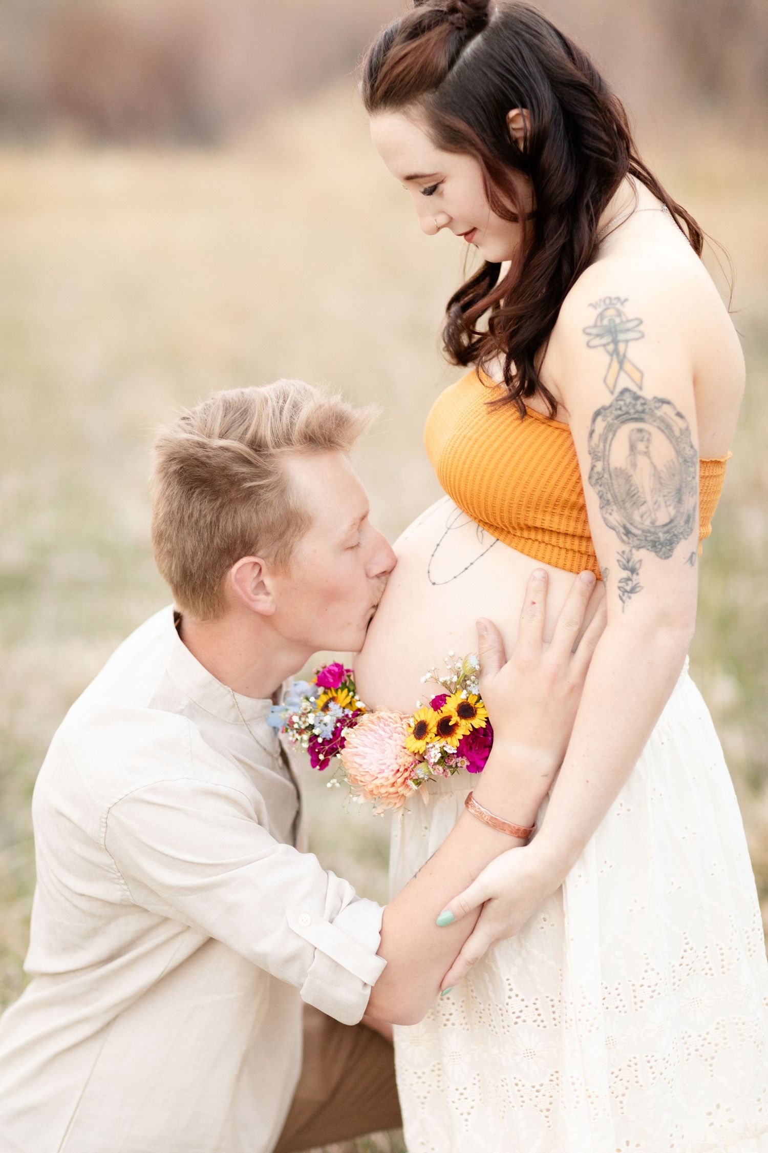 Man kneeling to kiss pregnant woman's belly while she smiles down at him in a golden field.
