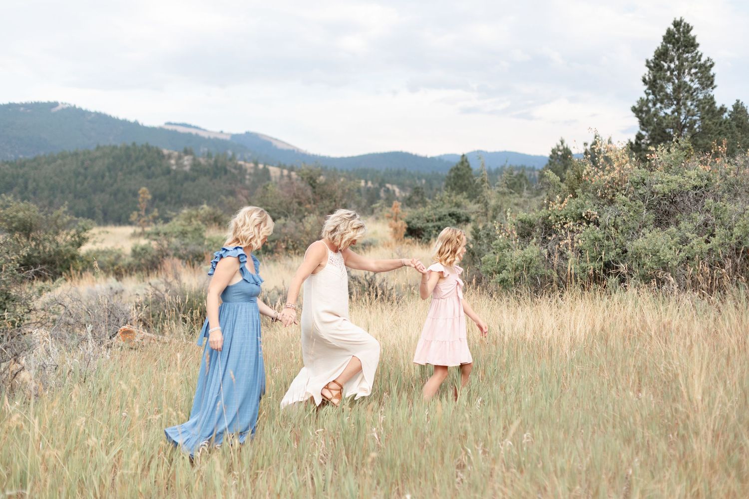 Woman in blue dress and girl in pink dress dancing with a child in a mountain meadow.
