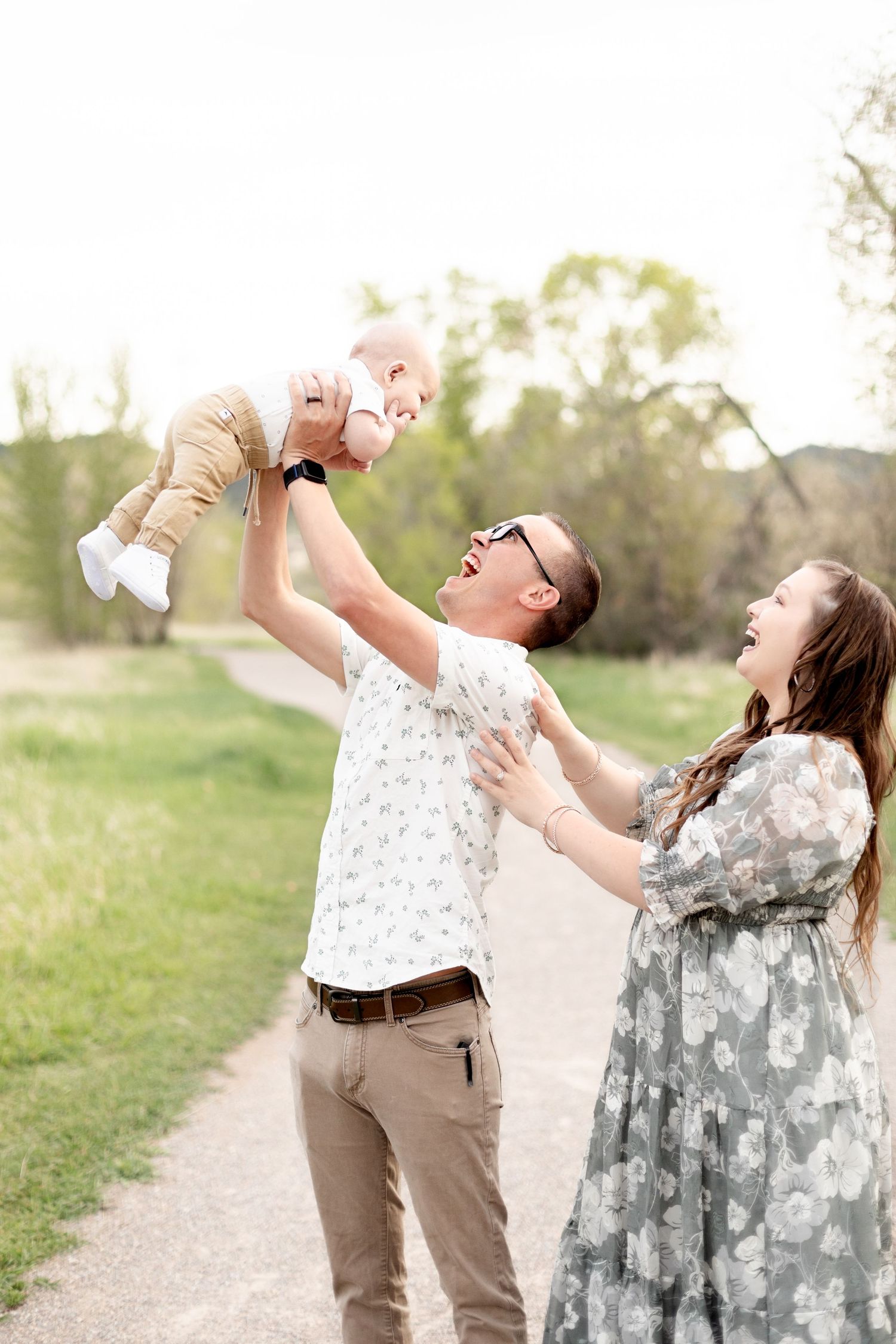 Father lifting laughing baby in the air while mother reaches up on a park path.