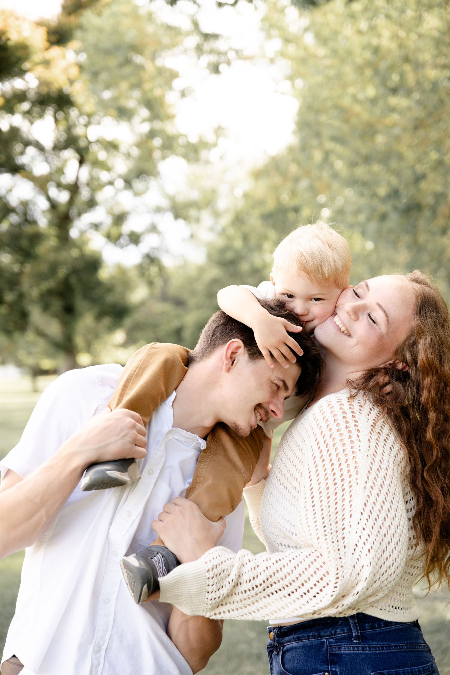 Joyful family moment as toddler sits on dads shoulders while mom leans in for a kiss outdoors.