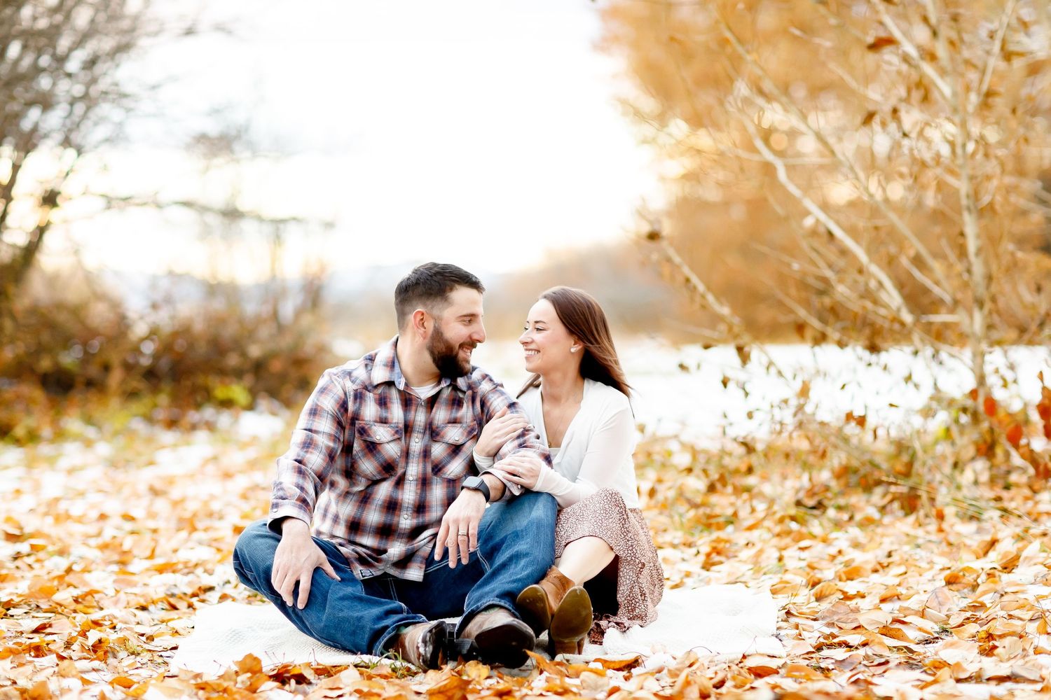 Couple sitting together on fallen autumn leaves, smiling at each other outdoors.