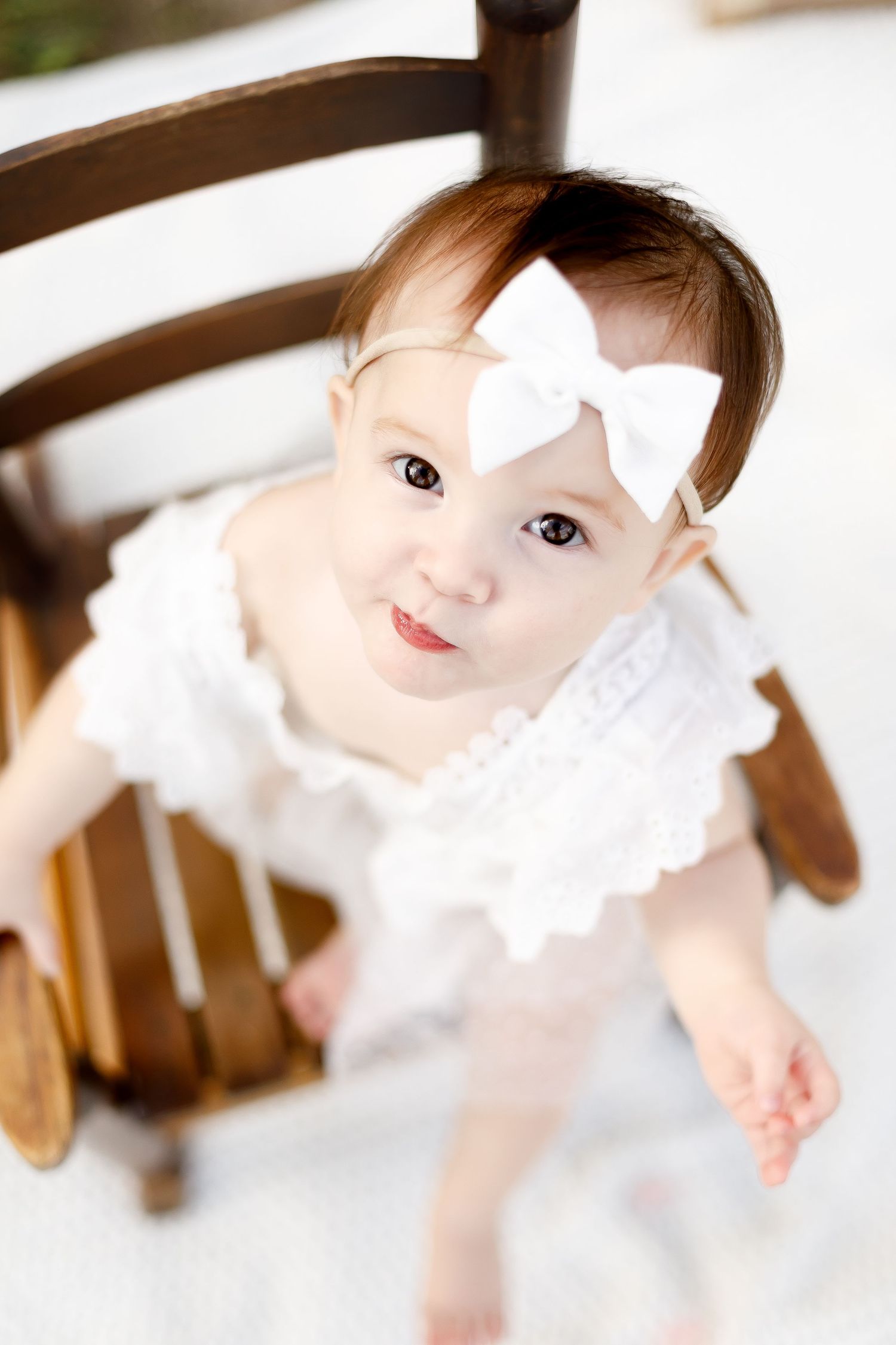Adorable baby girl in white lace romper with bow headband sitting on vintage wooden rocking chair.