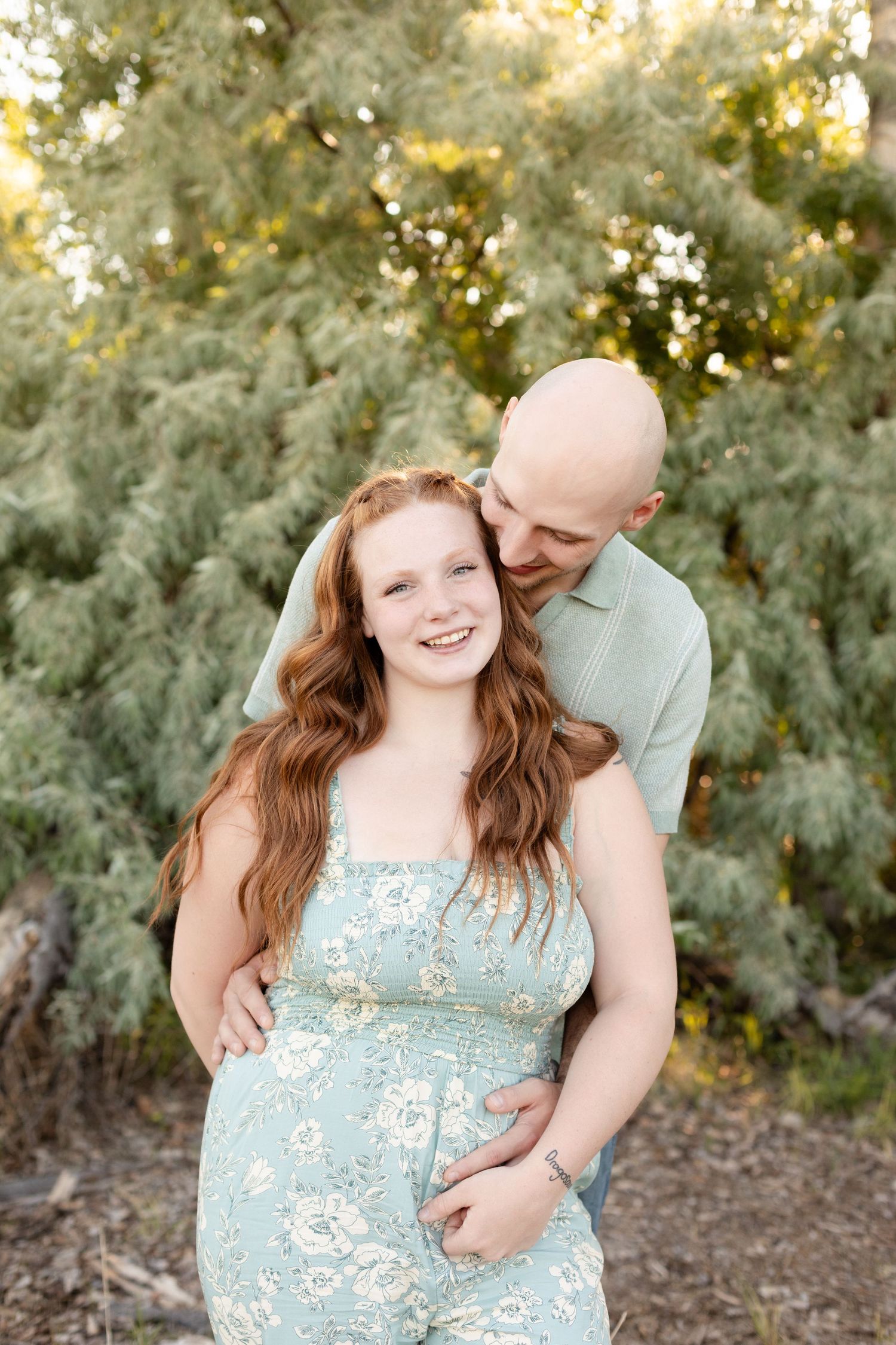 Smiling redhead woman in floral dress with bald man embracing her outdoors near green trees.