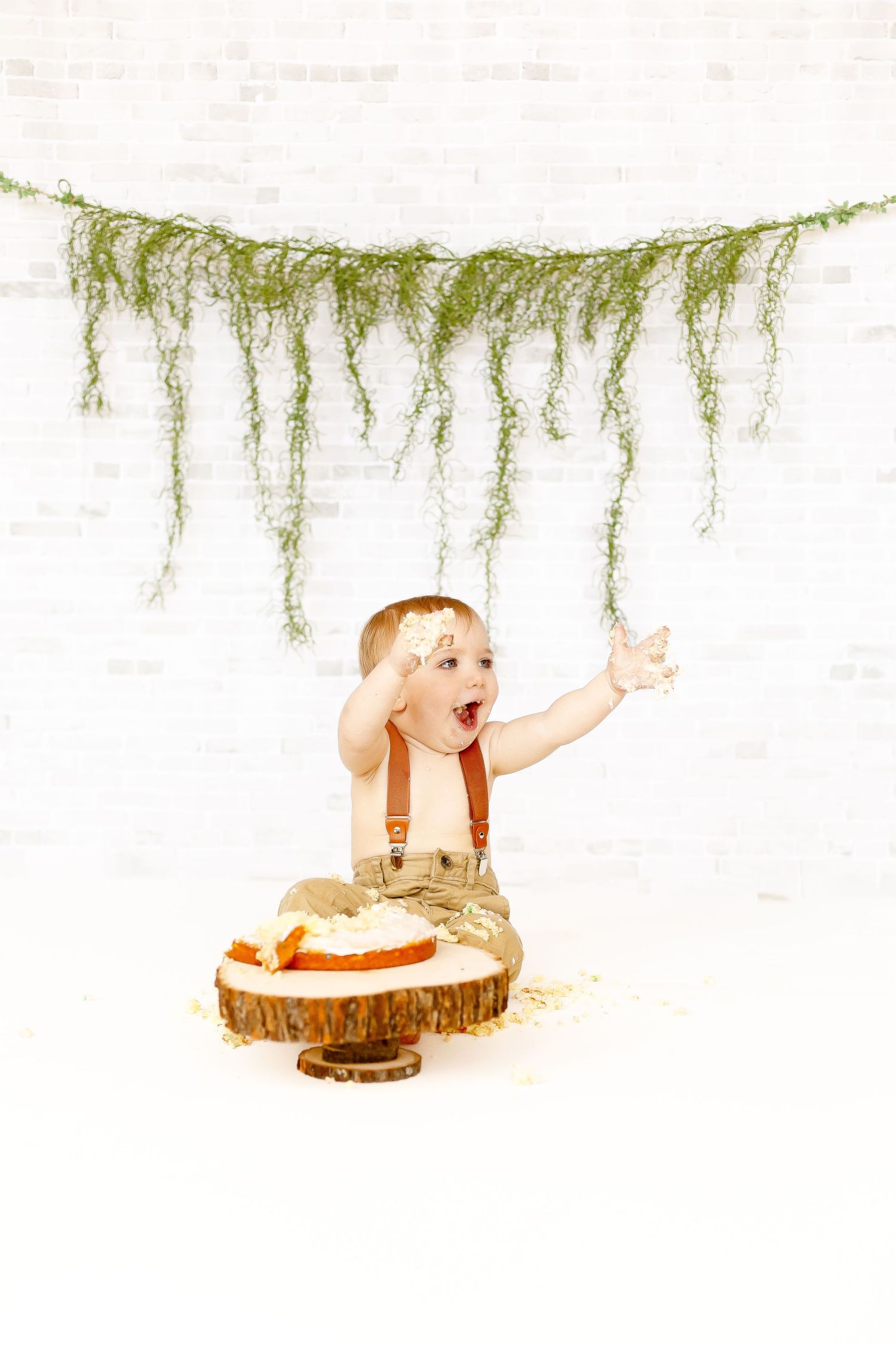 An excited baby boy with arms raised during a cake smash birthday photoshoot with greenery backdrop.