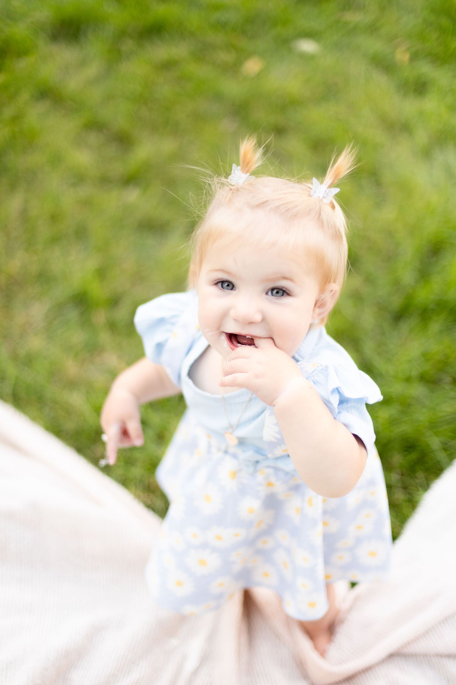 Adorable blonde baby girl in a blue daisy dress with pigtails smiling and reaching forward on a blanket outdoors.