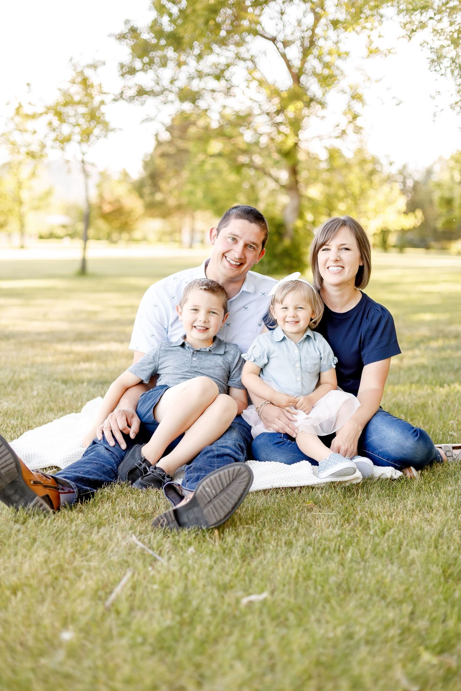 Smiling family of four sits together on a blanket in a sunny park.