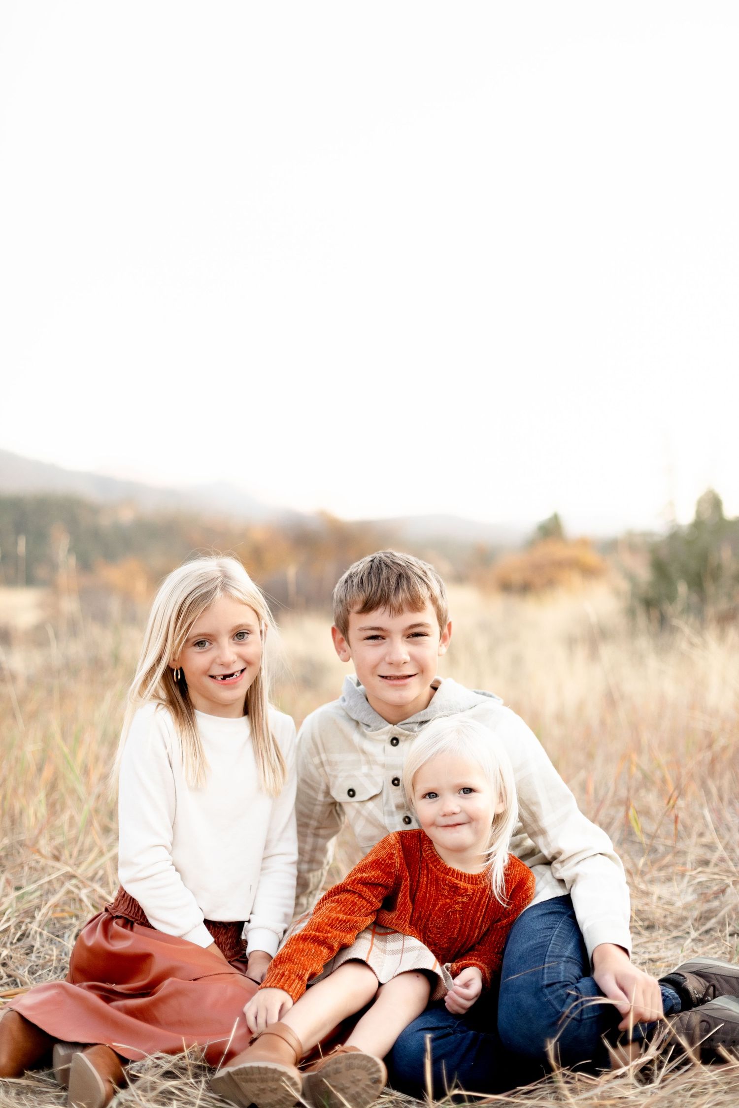Three siblings sitting together in a dry autumn field, smiling at the camera during fall portraits.