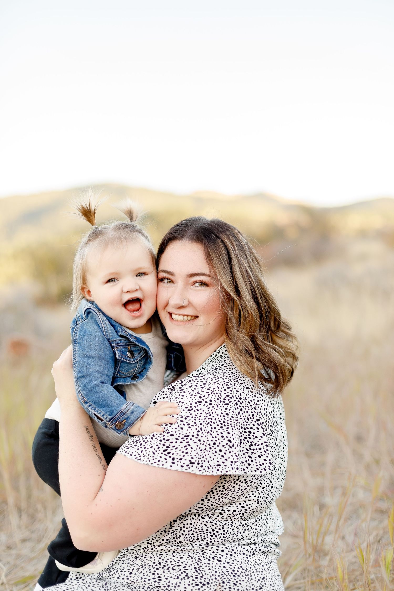 A smiling mother holds her laughing toddler girl in a denim jacket outdoors in a golden field.