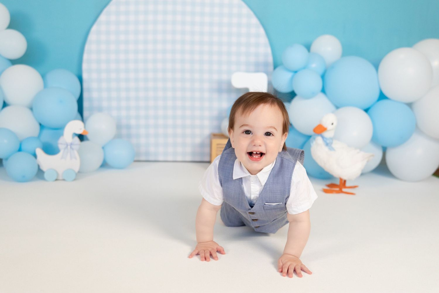 Smiling baby boy in a gray vest crawling on white floor with blue balloons and duck decorations.
