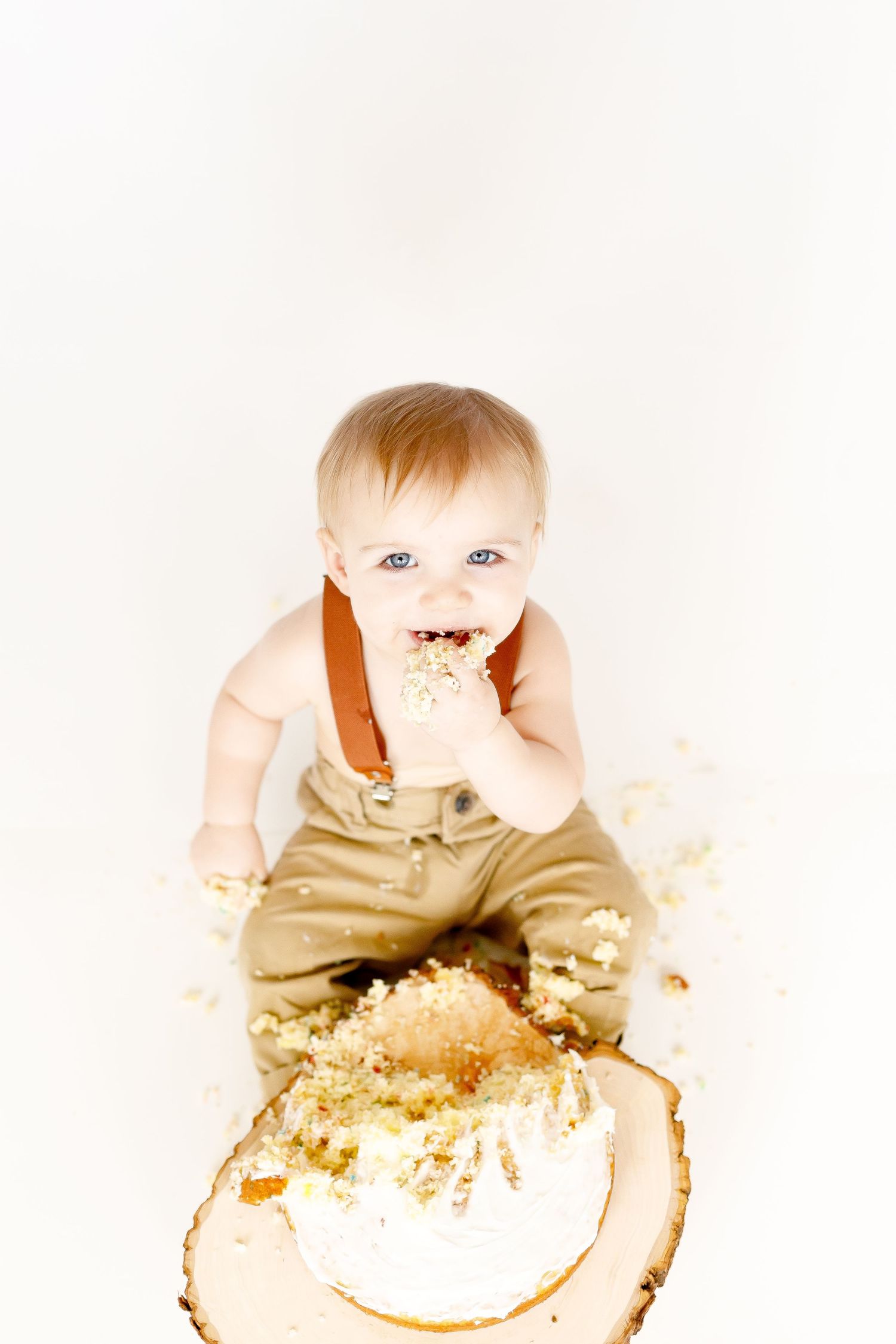 A joyful baby boy eating cake with his hands during a first birthday cake smash photoshoot.