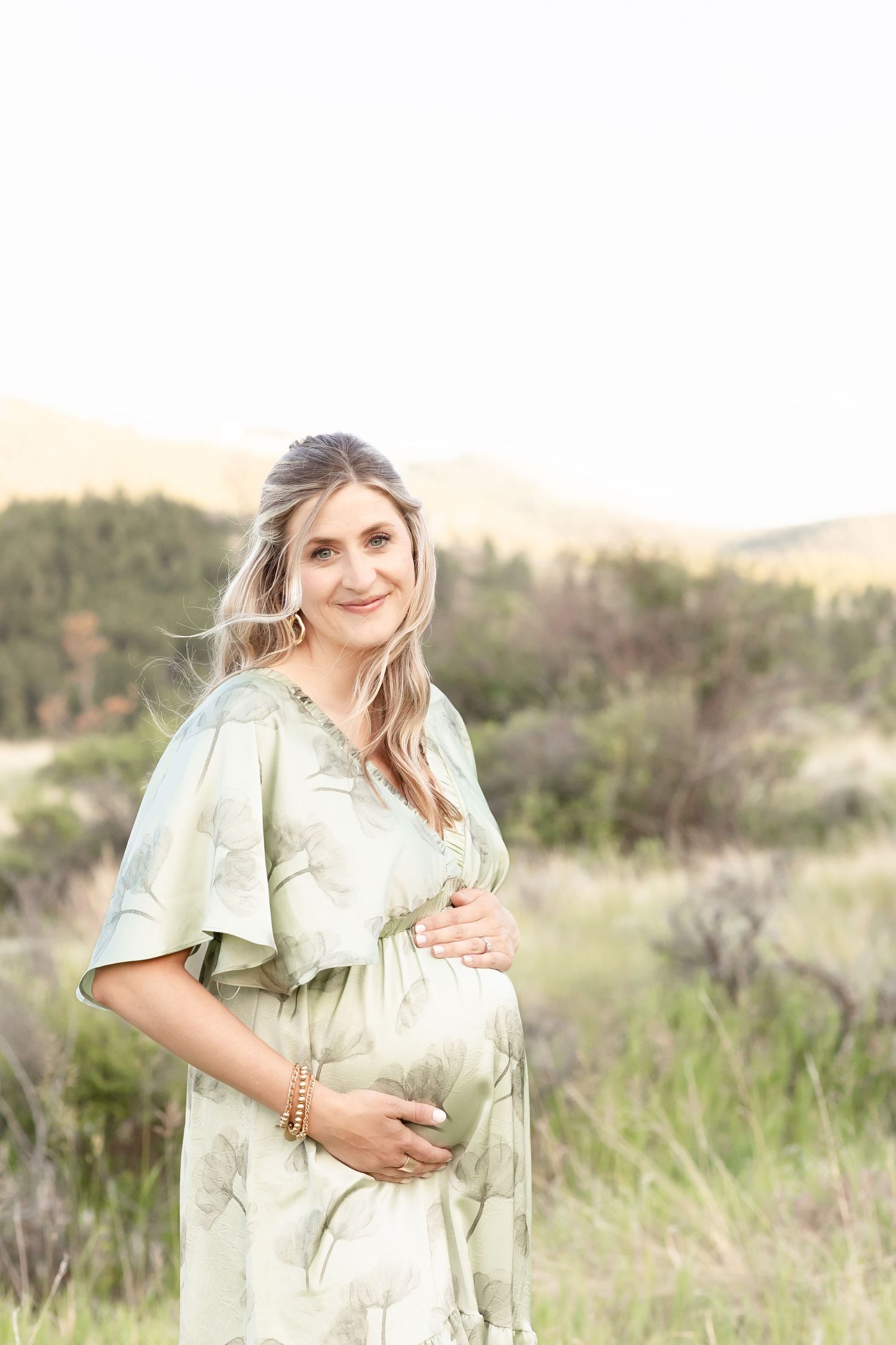 Smiling pregnant woman in sage green floral dress holding baby bump in scenic mountain field.