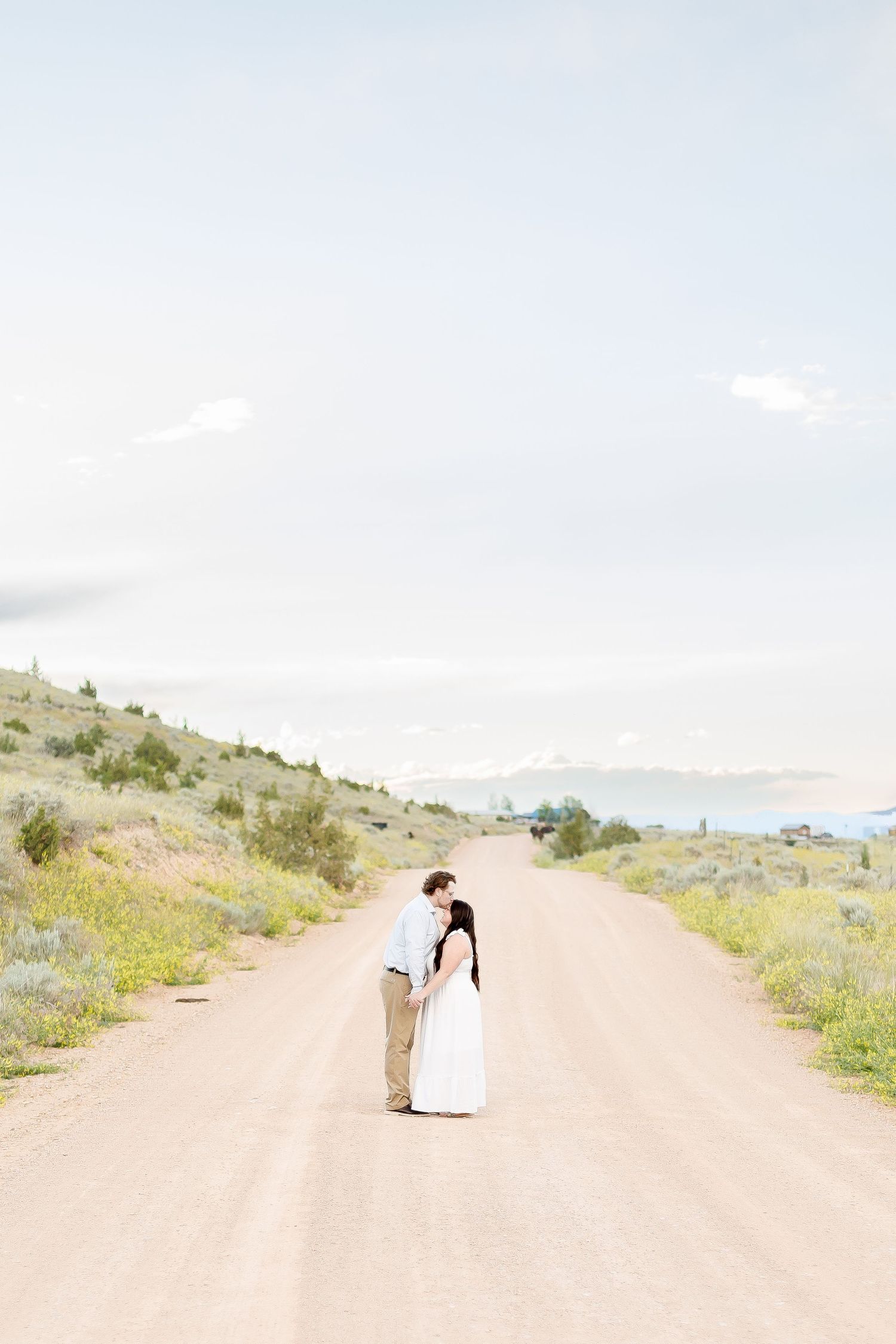 Couple sharing a romantic kiss on a long dirt road with mountain views in the background.