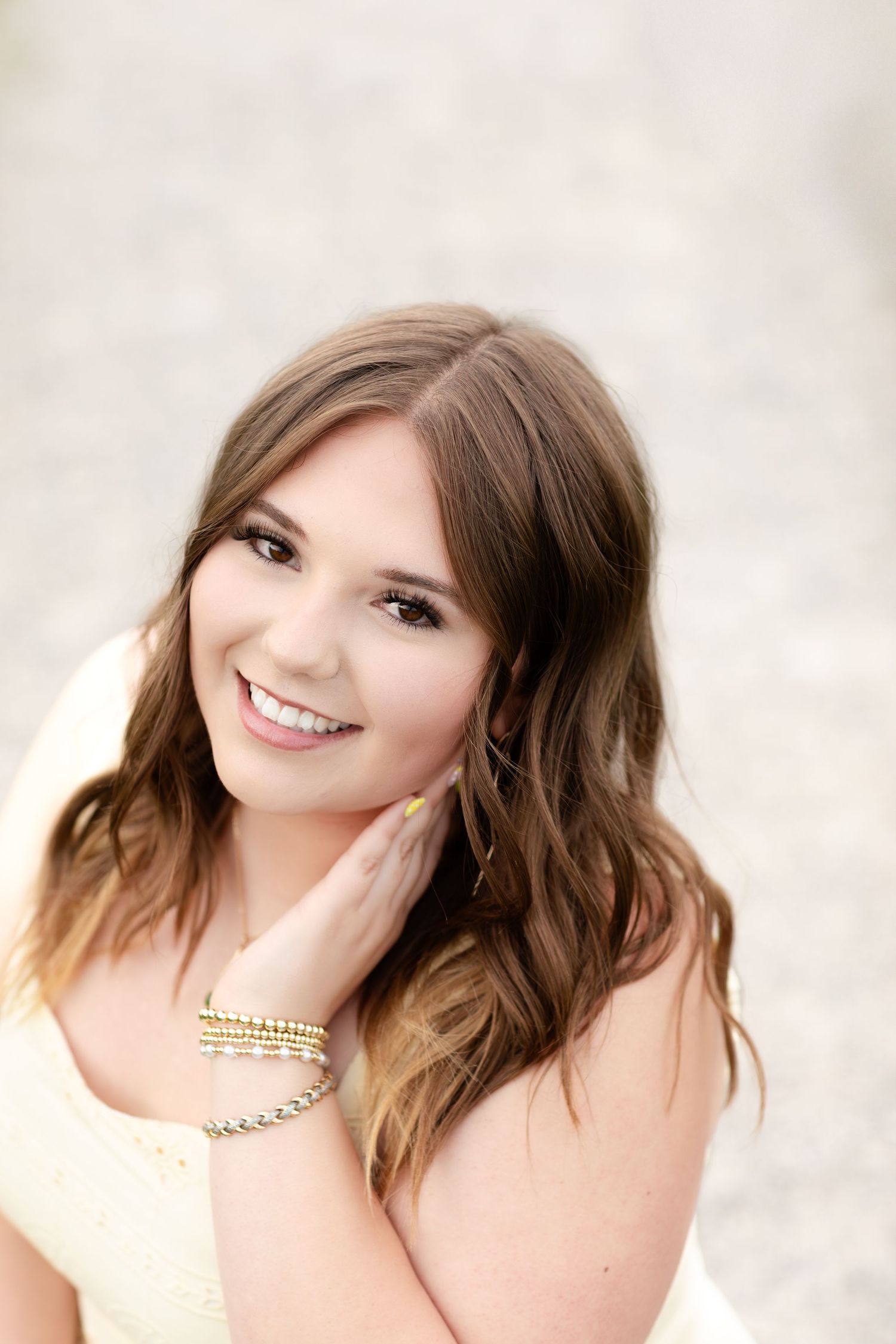 Smiling young woman with wavy brown hair touching her face wearing bracelets and a light yellow top.