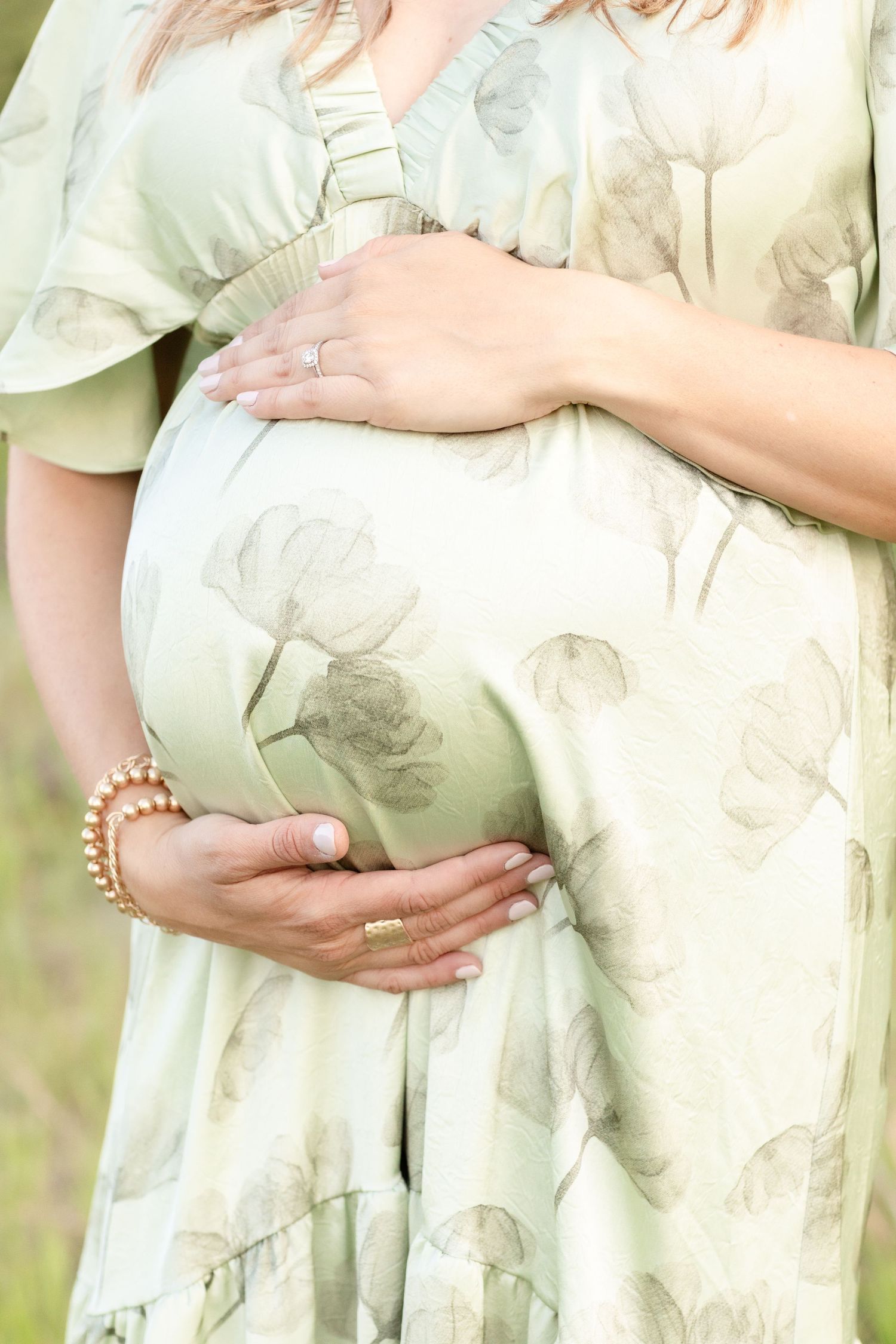 Close-up of pregnant woman in floral green dress cradling baby bump with manicured hands.
