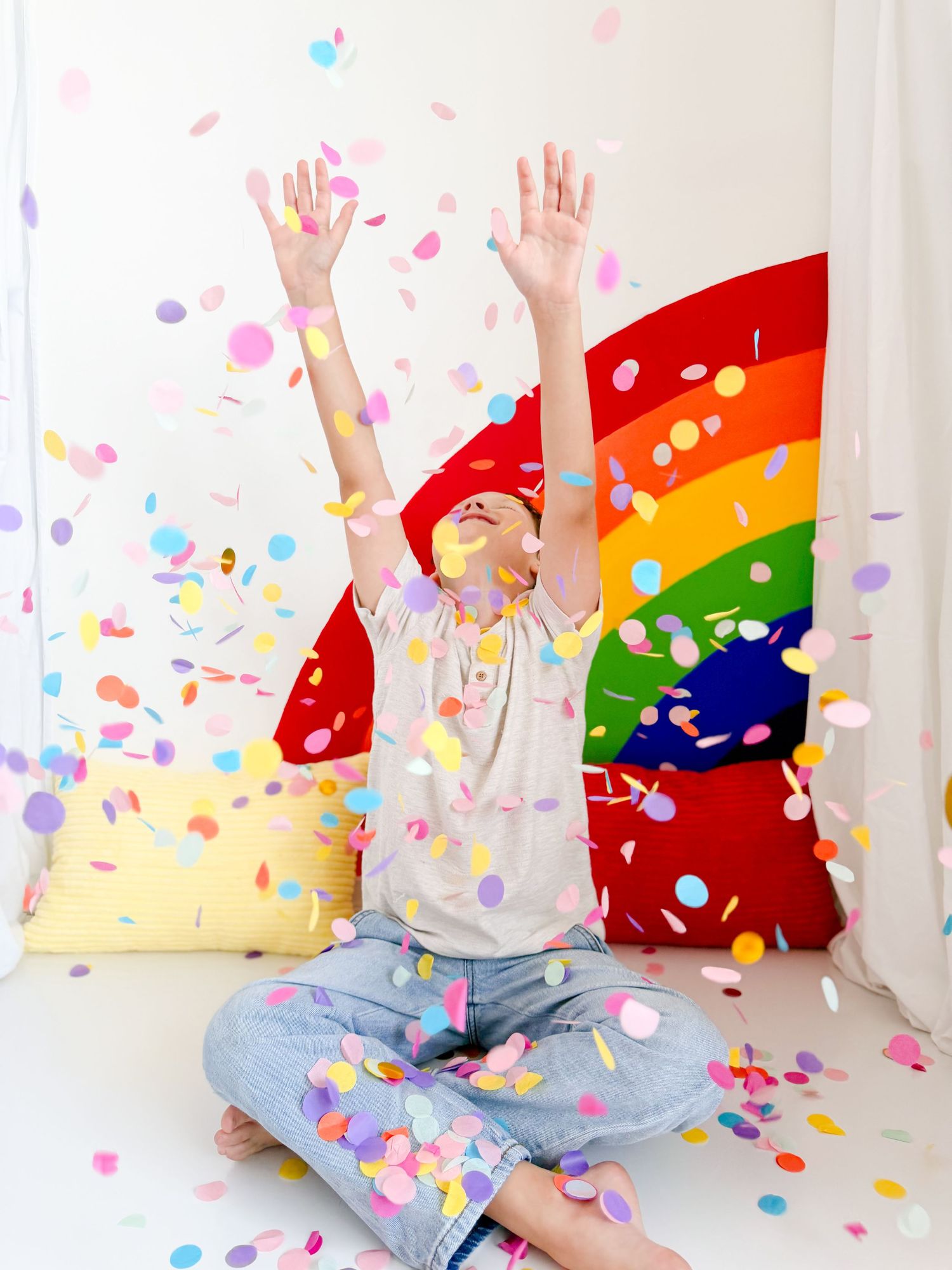 Child sitting cross-legged with arms raised joyfully, surrounded by colorful confetti falling near a rainbow backdrop.
