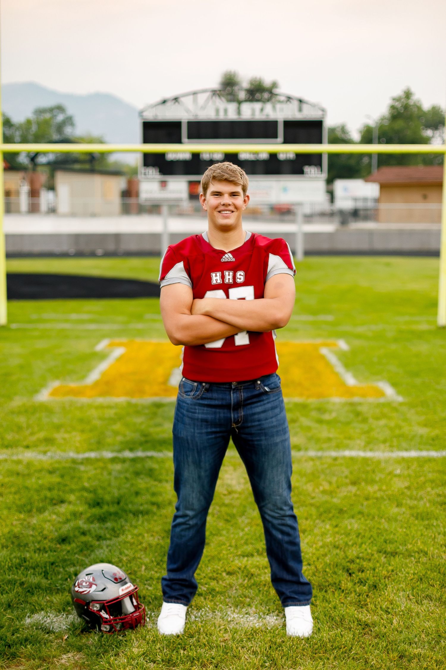 Teen in HHS red football jersey standing on football field with arms crossed and helmet at feet.