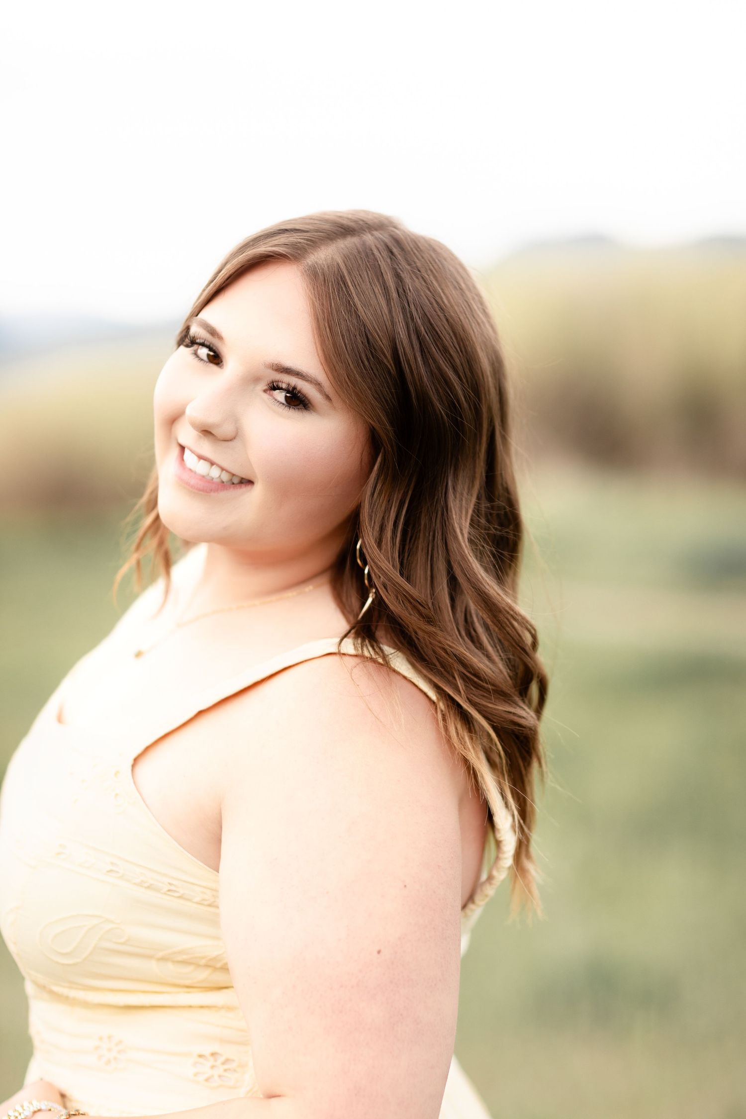 Smiling young woman in yellow dress with wavy brown hair posing outdoors in a bright open field.
