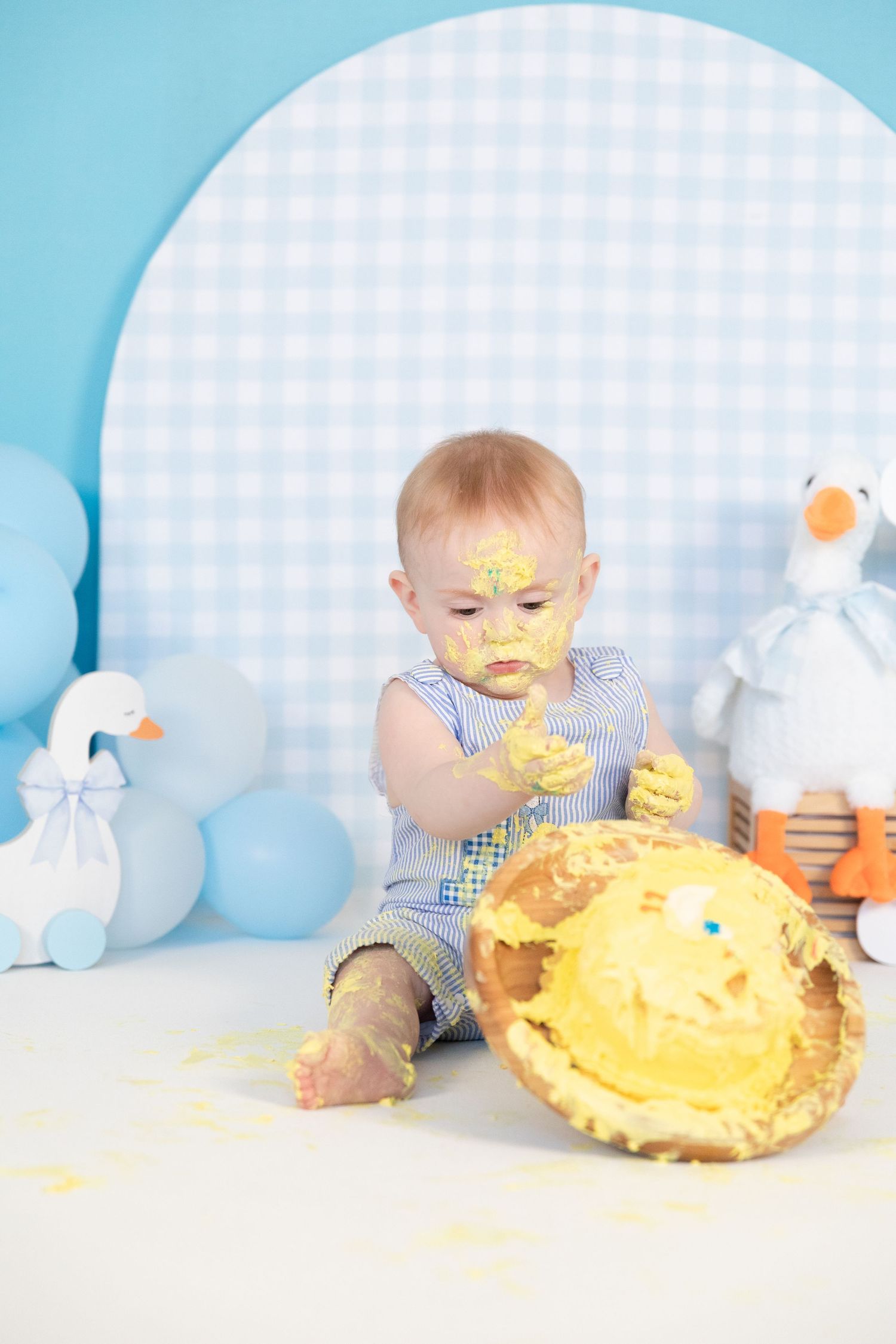 Baby boy with cake-covered face smashing yellow birthday cake during first birthday photo session.