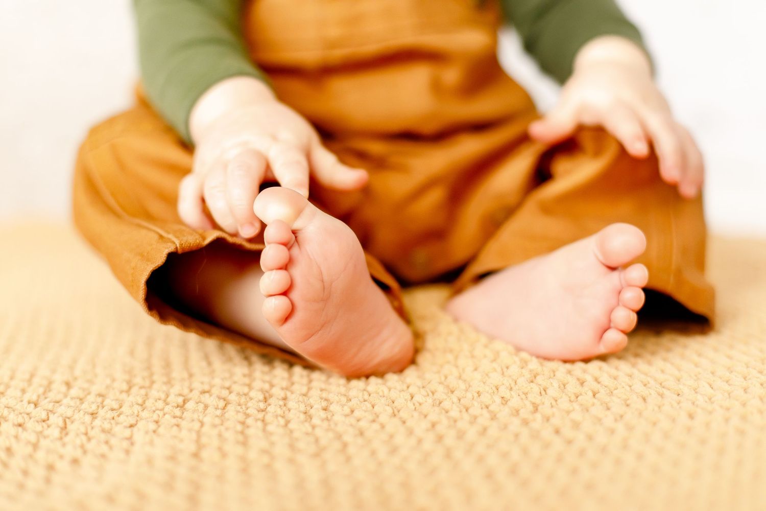 Close-up of baby feet with tiny hands touching toes while sitting on a knit blanket.