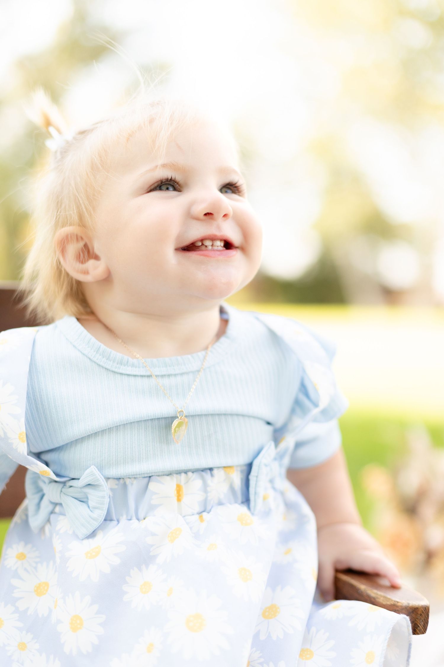 Happy blonde baby girl in a blue daisy dress laughing and looking up while sitting outdoors in sunlight.
